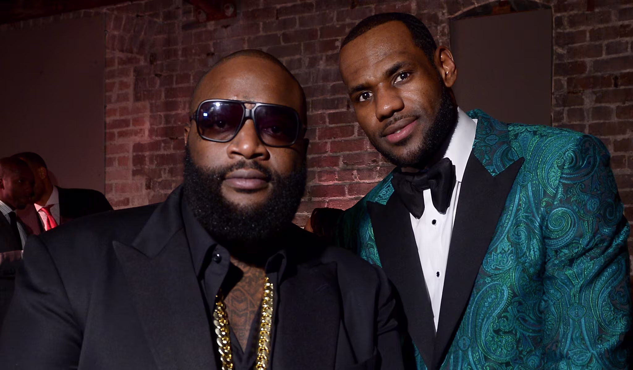 Rick Ross and LeBron James posing together. Rick Ross wears sunglasses and a black suit, LeBron in a teal patterned tuxedo.