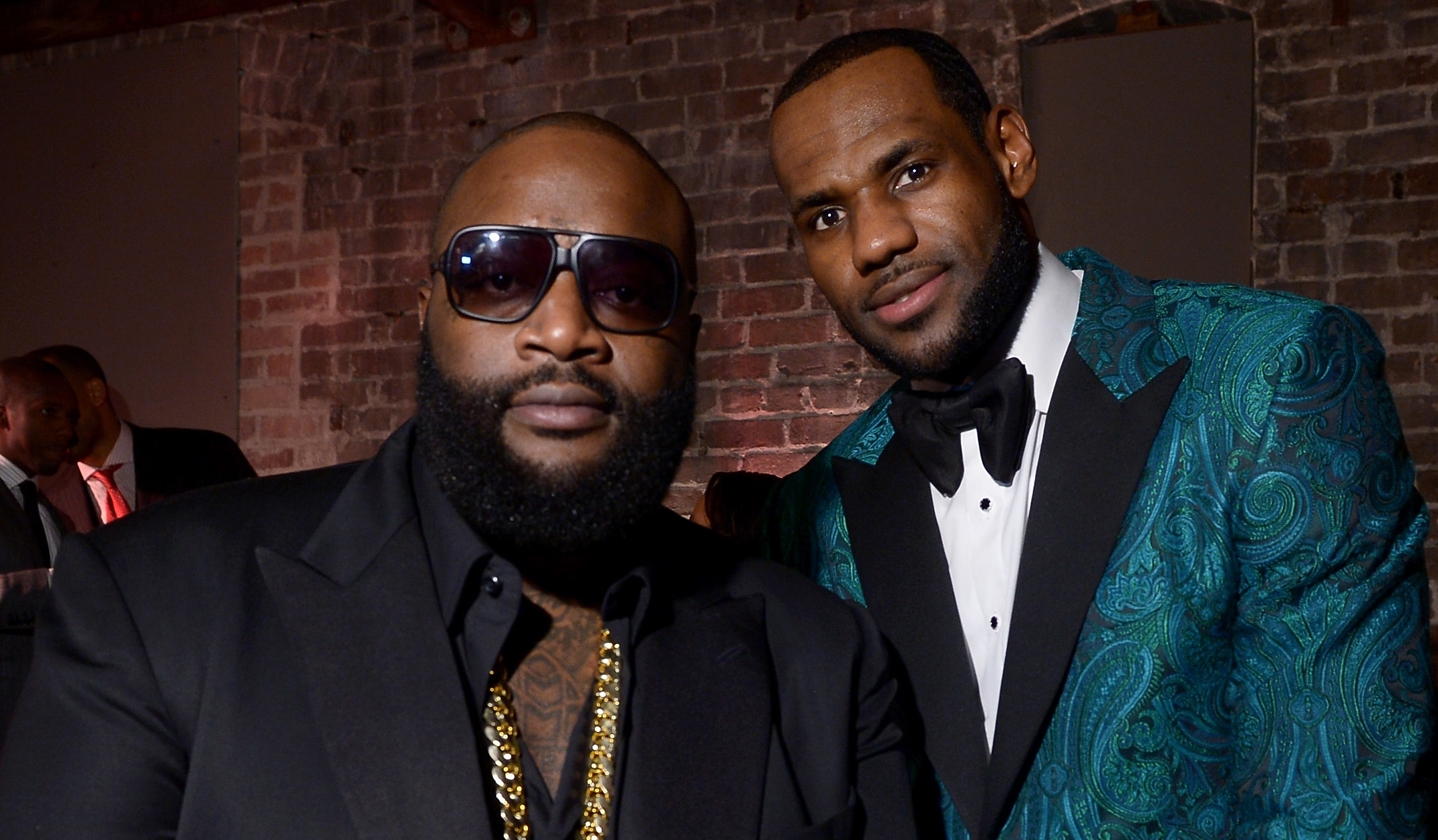 Rick Ross and LeBron James posing together. Rick Ross wears sunglasses and a black suit, LeBron in a teal patterned tuxedo.