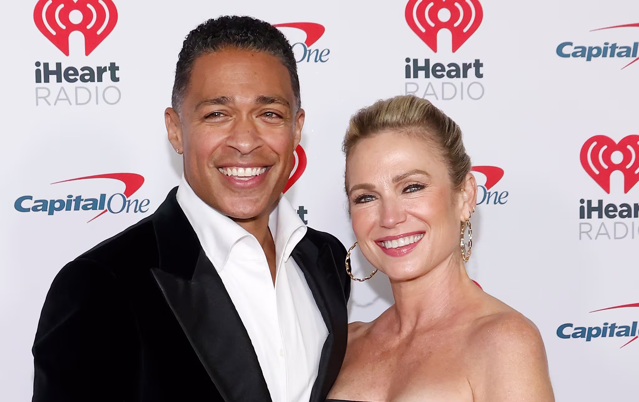 T.J. Holmes in a black suit and Amy Robach in a black dress pose together, smiling at an iHeartRadio event.