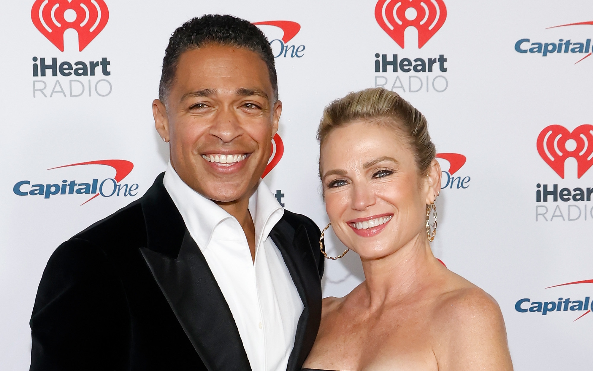 T.J. Holmes in a black suit and Amy Robach in a black dress pose together, smiling at an iHeartRadio event.
