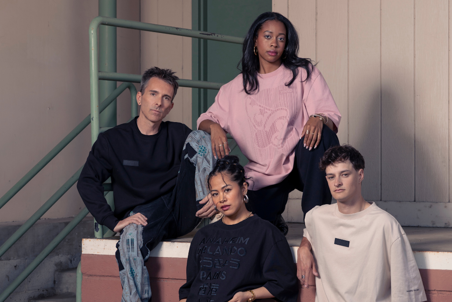 Four people pose fashionably on stairs, wearing casual oversized clothing in muted tones.