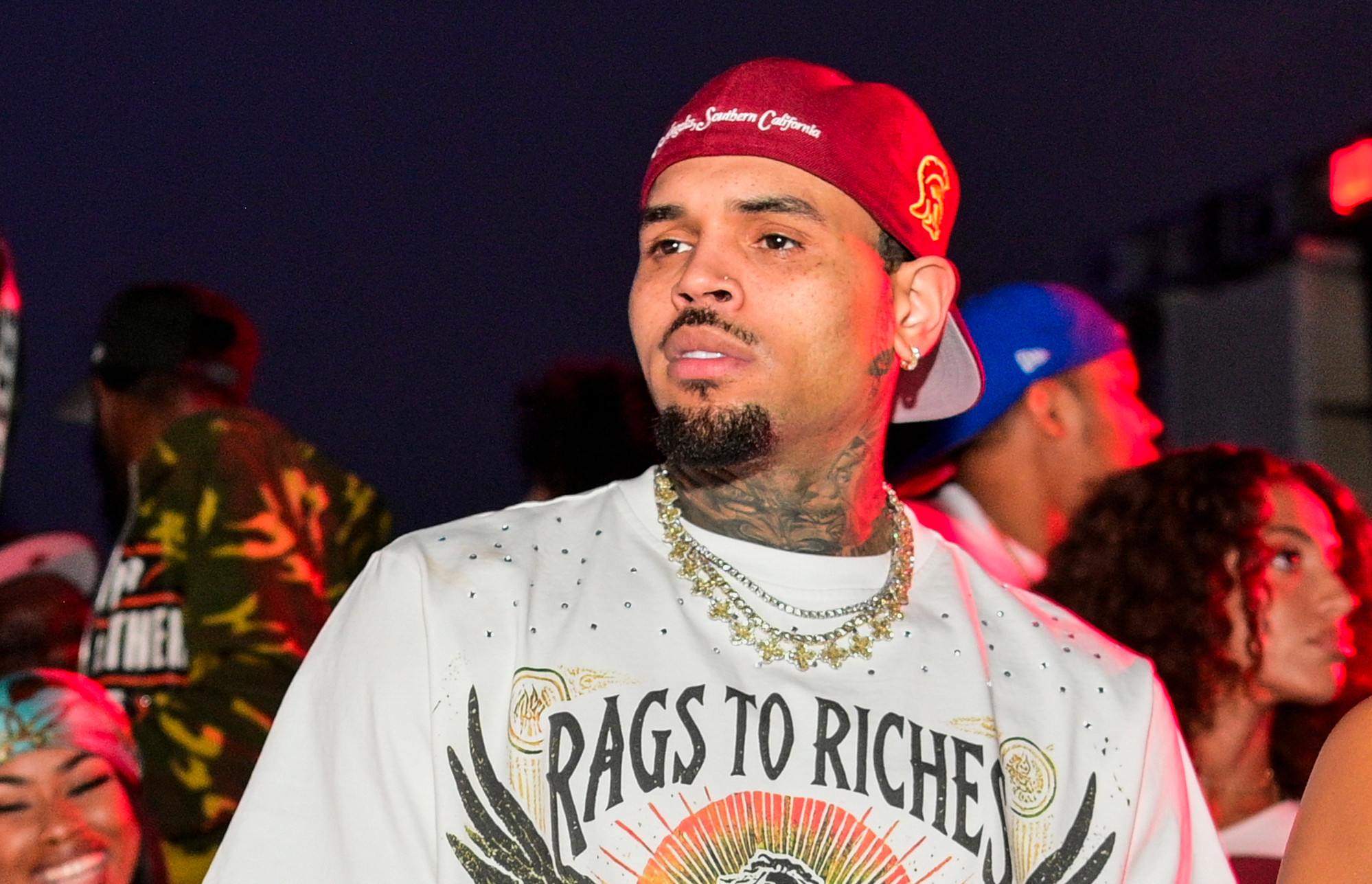 Chris Brown wearing a "Rags to Riches" shirt and a red cap stands in a lively crowd, with people enjoying the event around him.