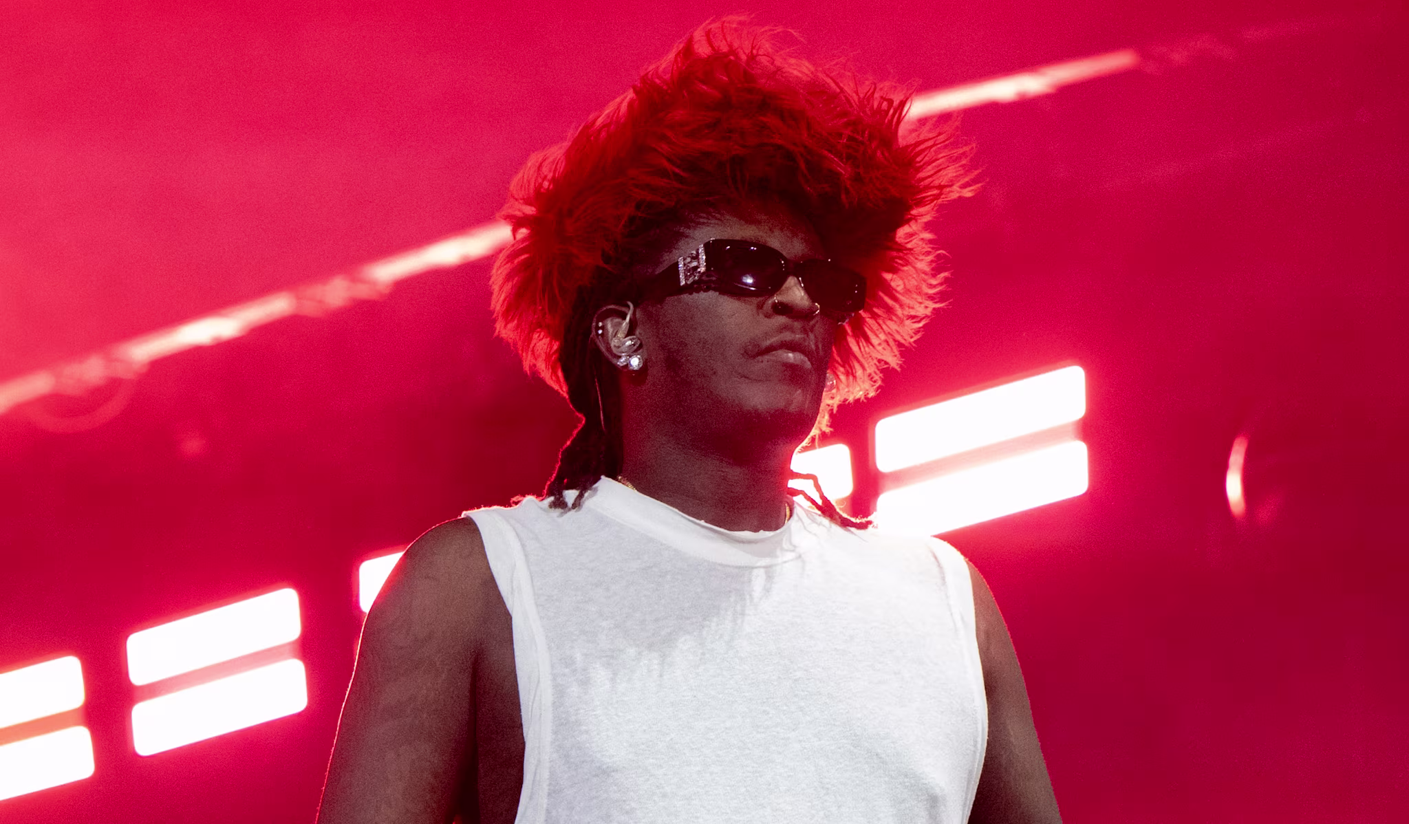 Young Thug on stage in a white sleeveless shirt, wearing sunglasses and a red feathered hat, with a microphone in hand.