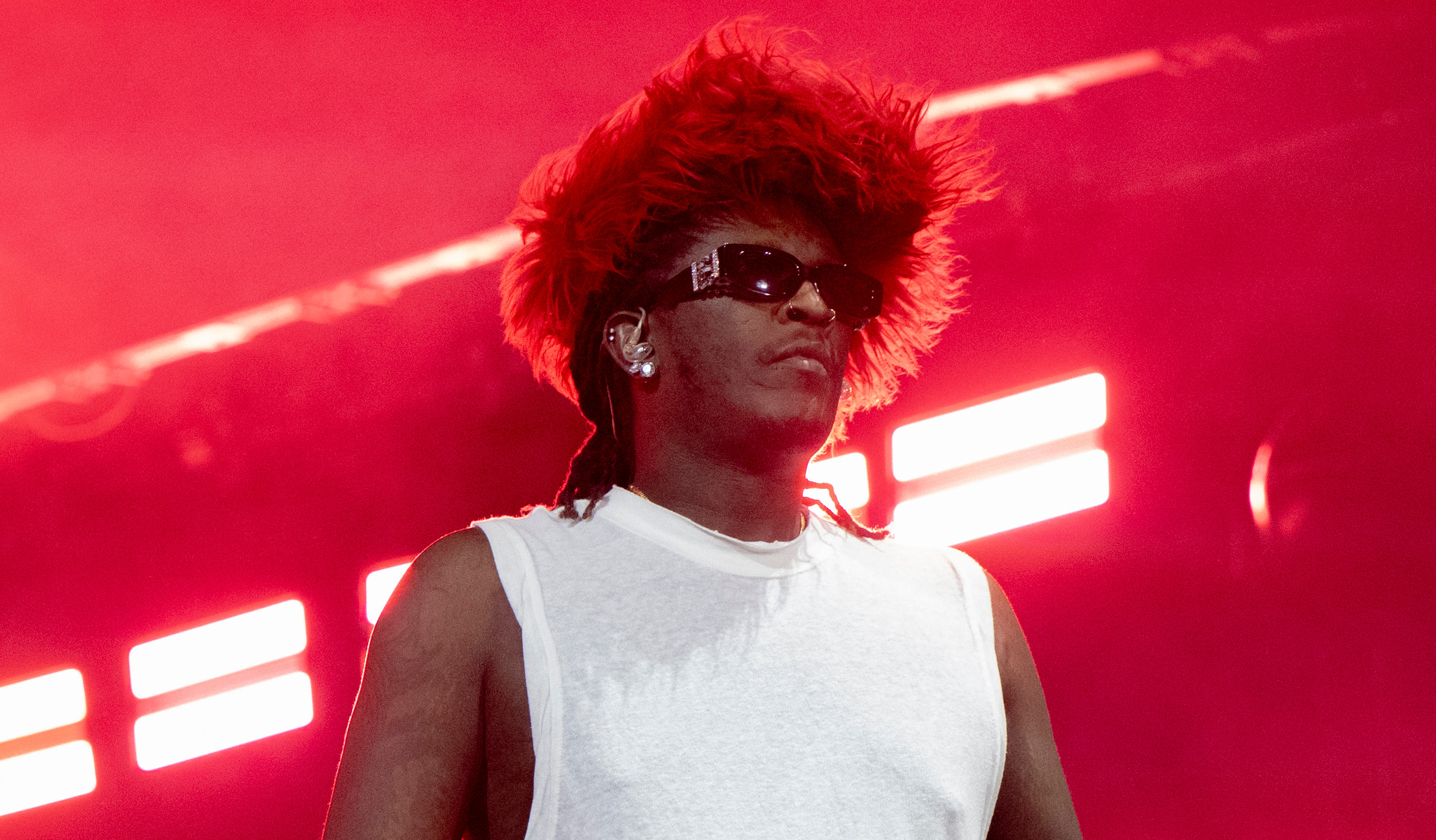 Young Thug on stage in a white sleeveless shirt, wearing sunglasses and a red feathered hat, with a microphone in hand.