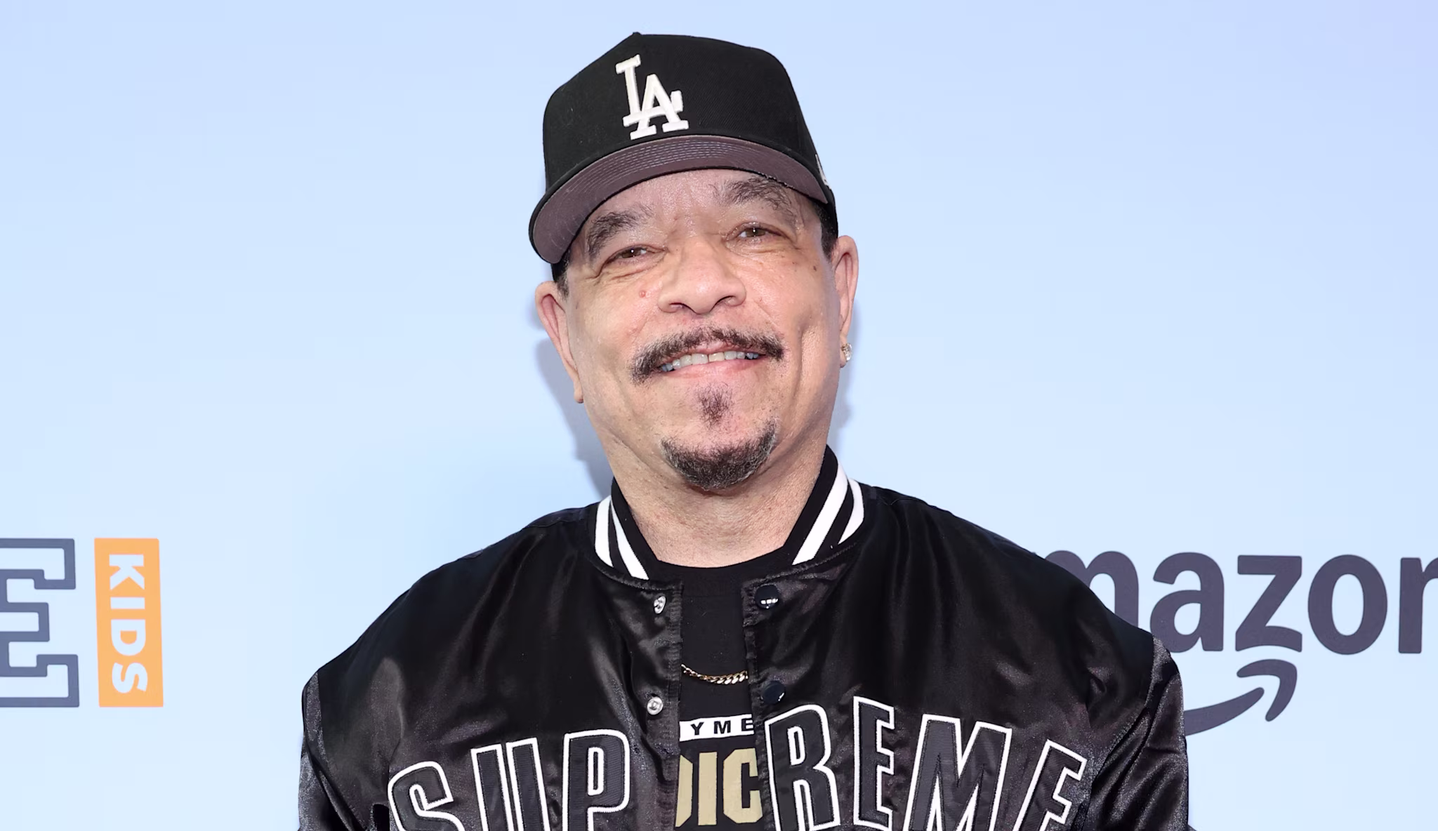 Ice-T in a Supreme jacket and LA cap on a red carpet, posing with peace signs. Background features brand logos.