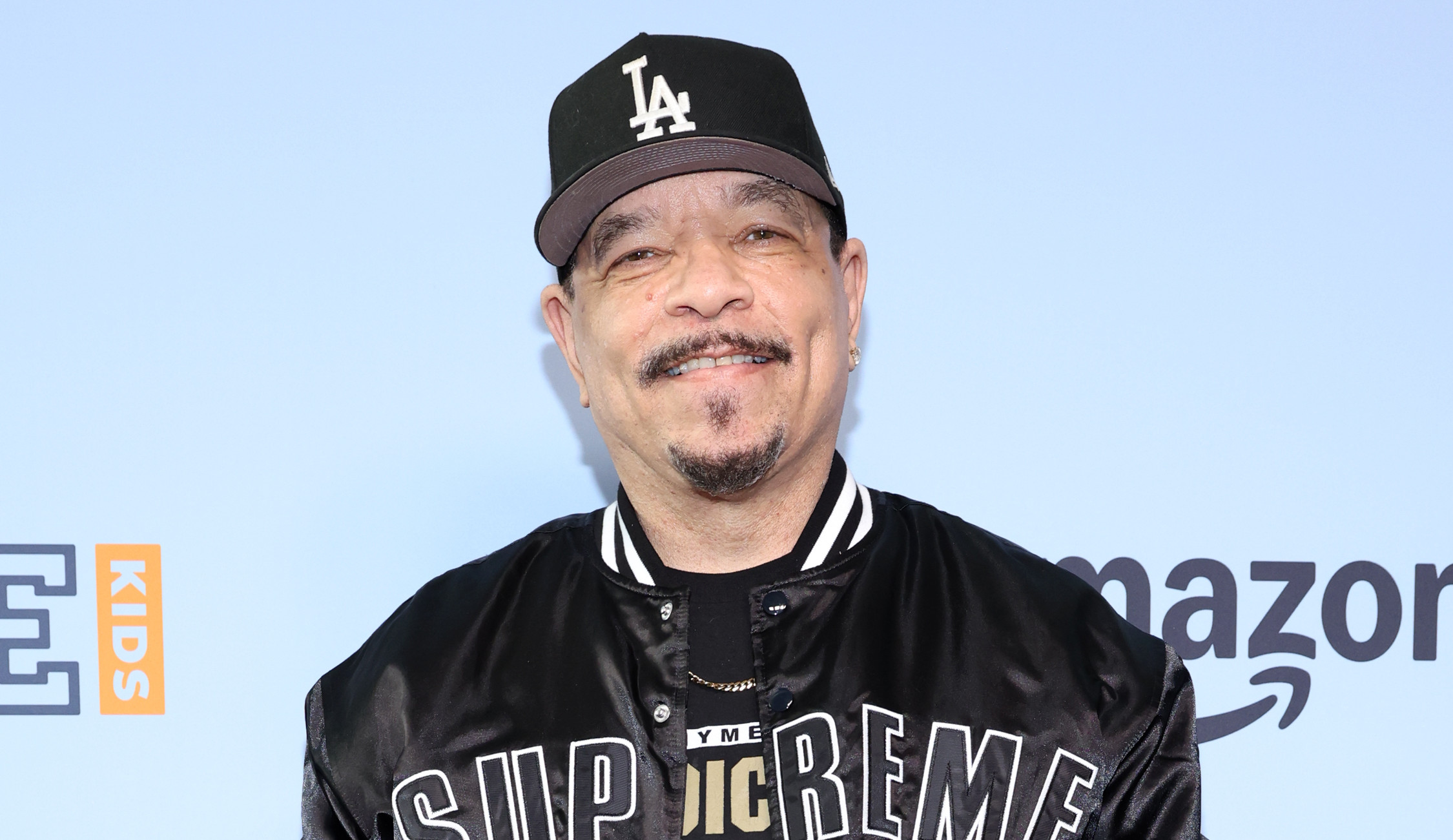 Ice-T in a Supreme jacket and LA cap on a red carpet, posing with peace signs. Background features brand logos.