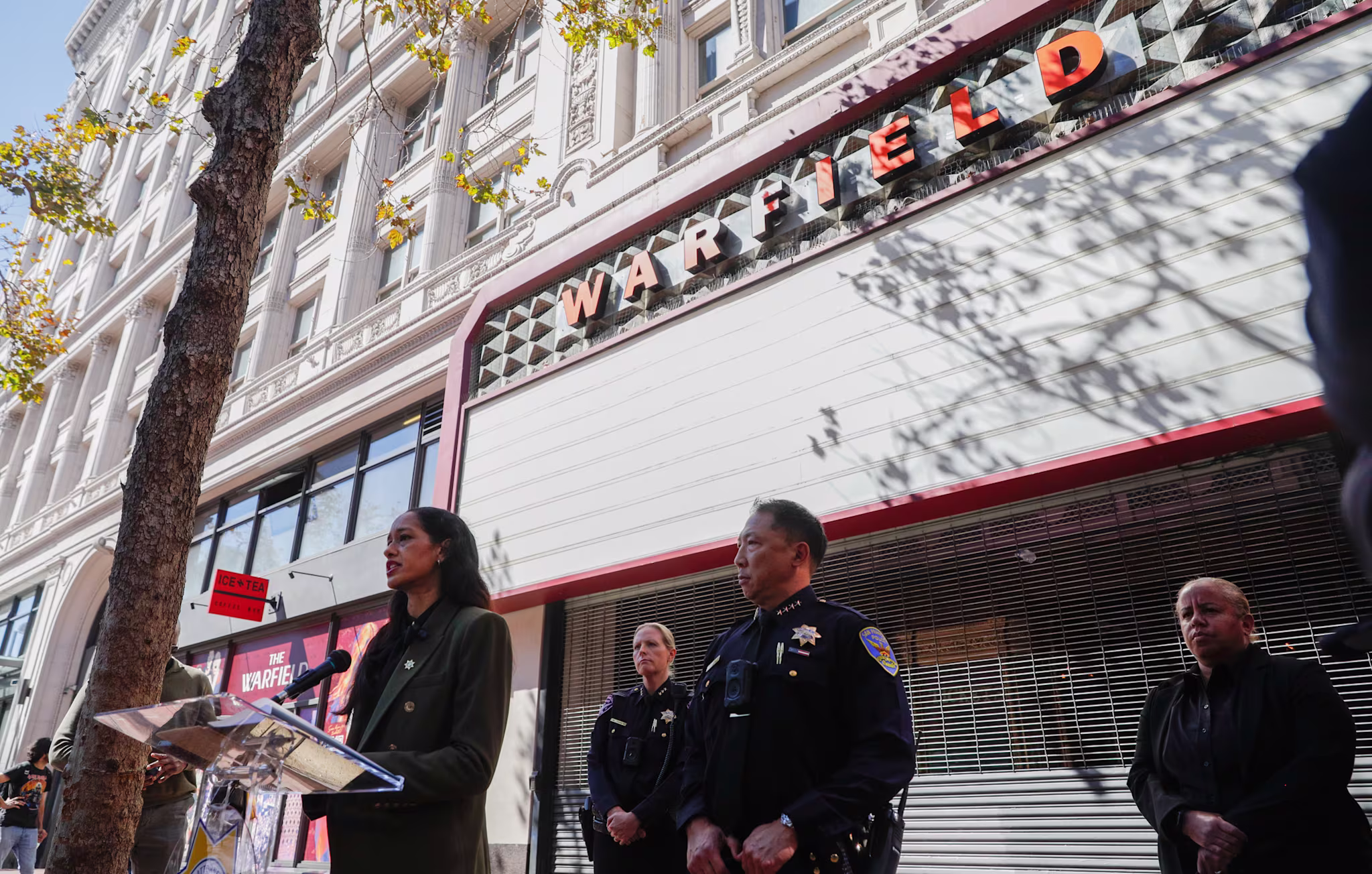 A person speaks at a podium outside the Warfield theater, surrounded by police officers and others, with a tree and building in the background.