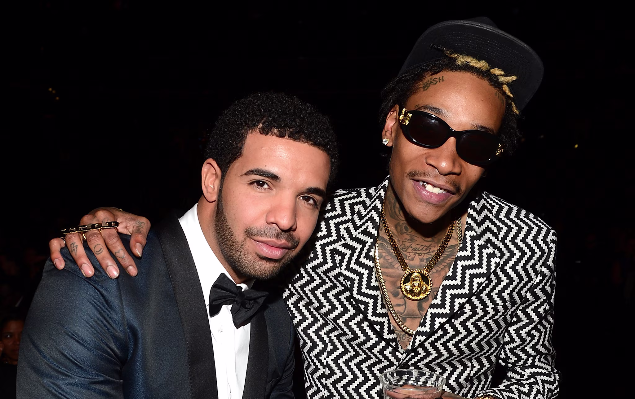 Drake in a dark suit and bow tie poses with Wiz Khalifa in a patterned suit and sunglasses, both smiling at an event.