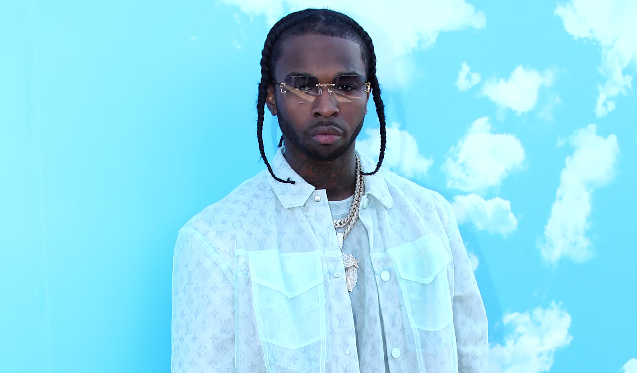 Pop Smoke in a light pastel outfit stands against a blue sky background with clouds, wearing sunglasses and white sneakers.
