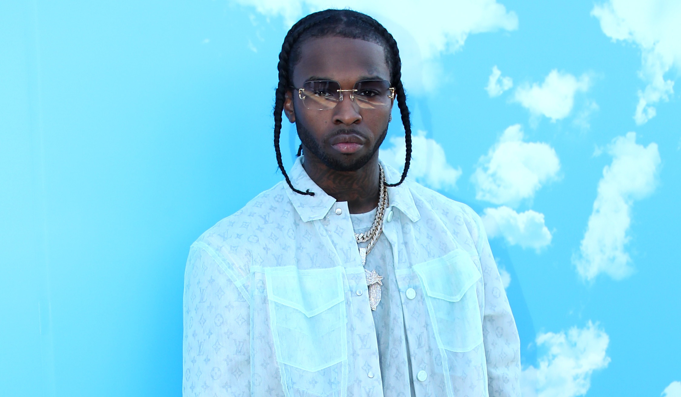 Pop Smoke in a light pastel outfit stands against a blue sky background with clouds, wearing sunglasses and white sneakers.