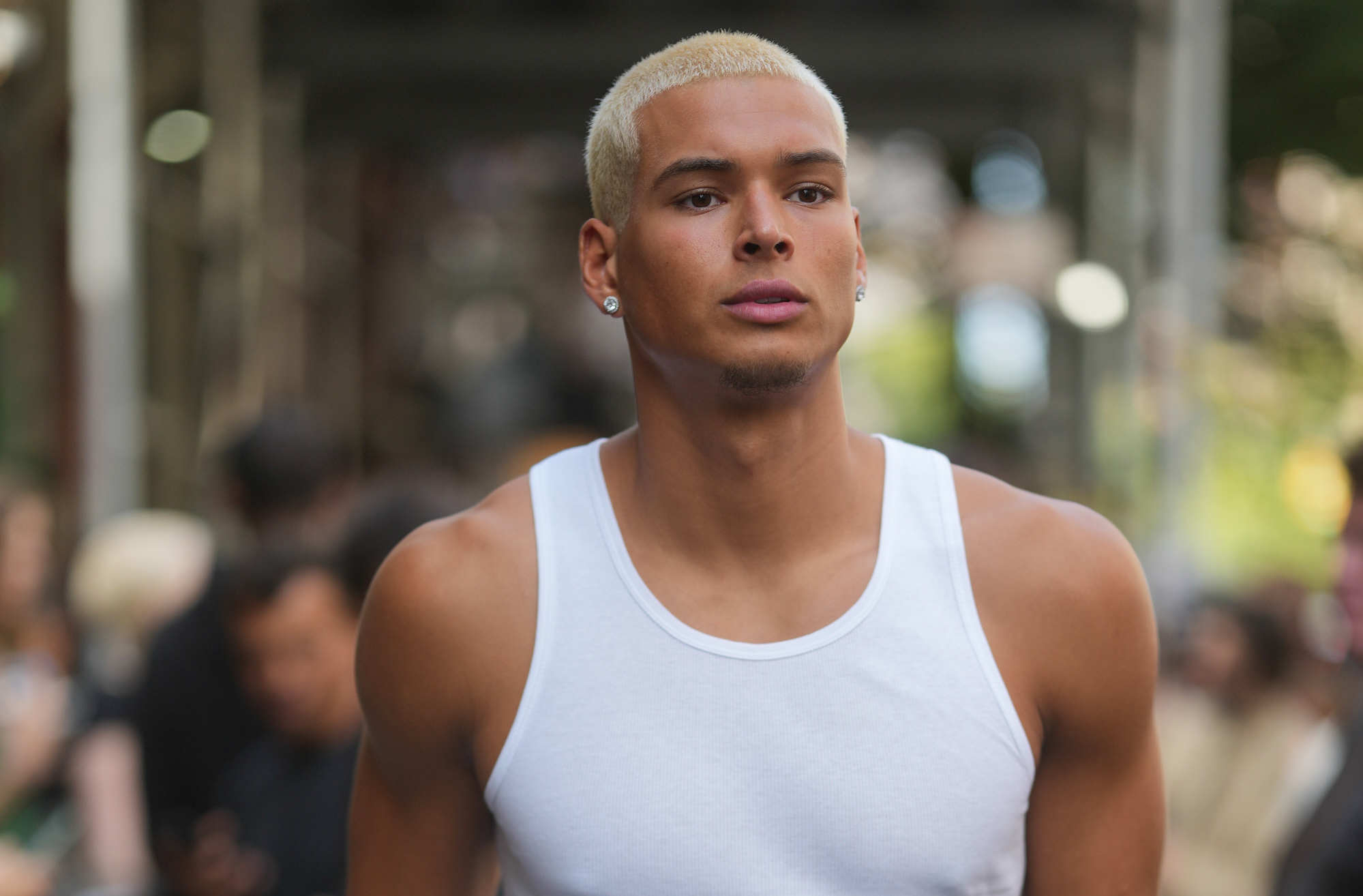 Marlon Lundgren Garcia with short blonde hair wearing a white tank top and black pants, holding a blue shirt, walks confidently on a city street.
