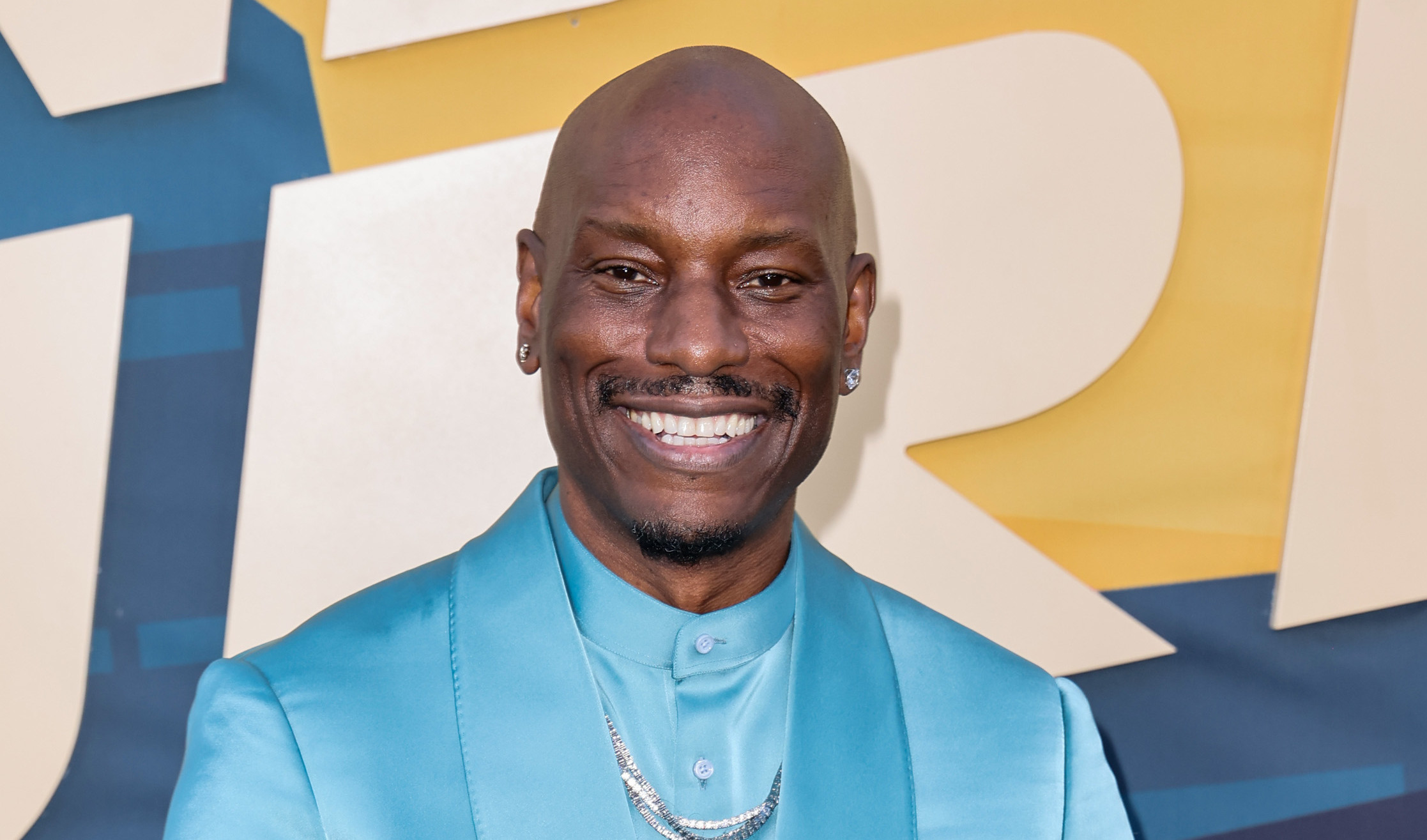 Tyrese Gibson in a shiny blue suit, smiling at the camera, standing in front of a colorful backdrop.