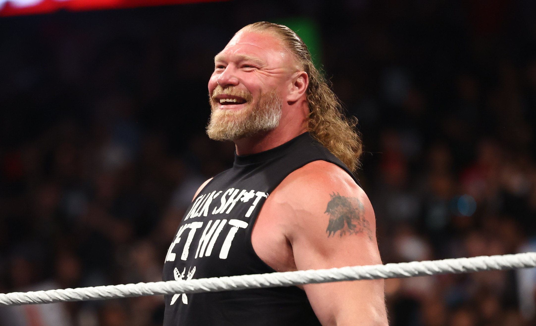 Brock Lesnar with long hair and a beard, wearing a sleeveless shirt, stands in a WWE ring, smiling at the audience.