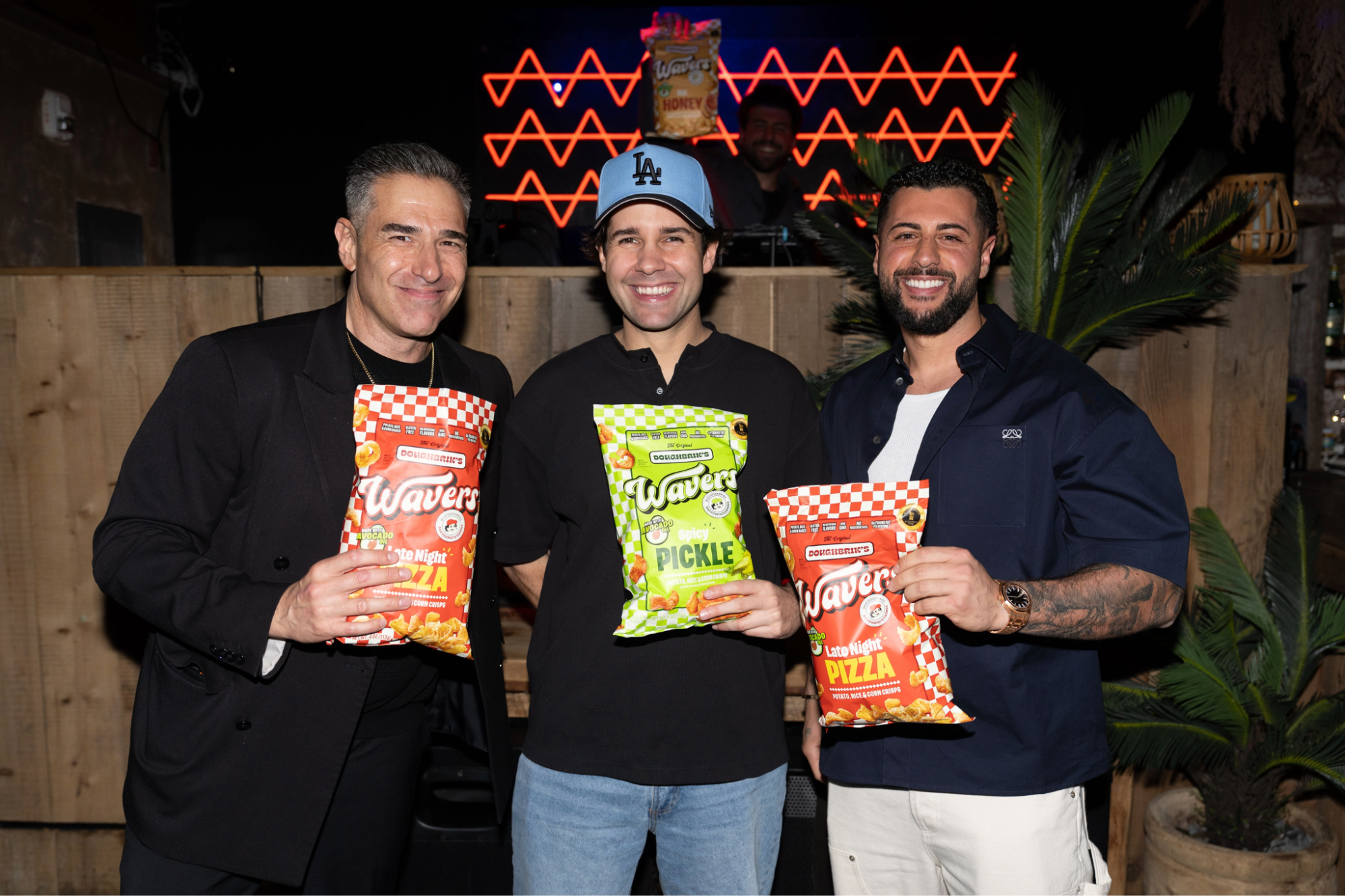 Three men smiling, each holding a bag of Wavy chips. They stand in front of a wooden backdrop with neon lights above.