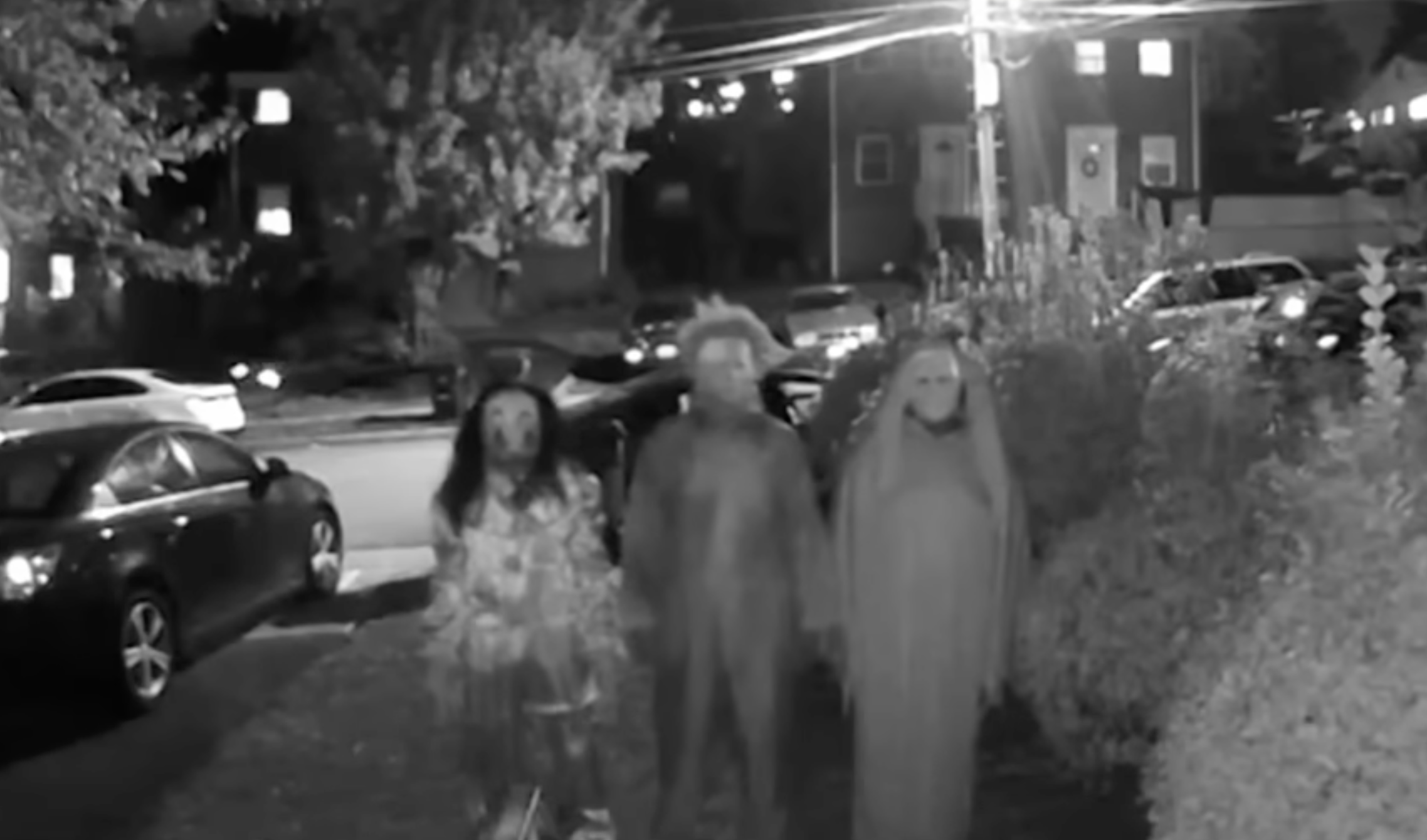 Three people in Halloween costumes, including a clown, a scarecrow, and a ghost, walk down a residential street at night.