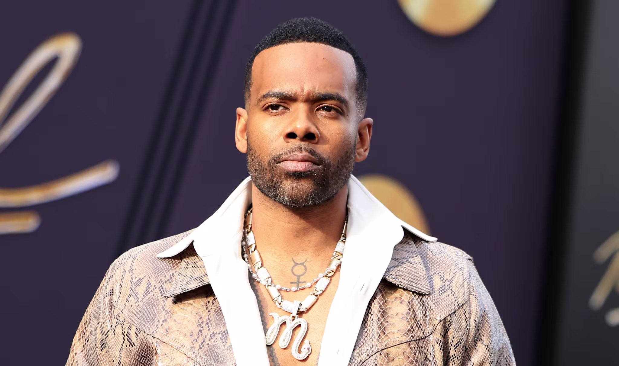 Mario in a snakeskin jacket and brown pants, wearing a necklace, stands in front of a dark background with gold accents.