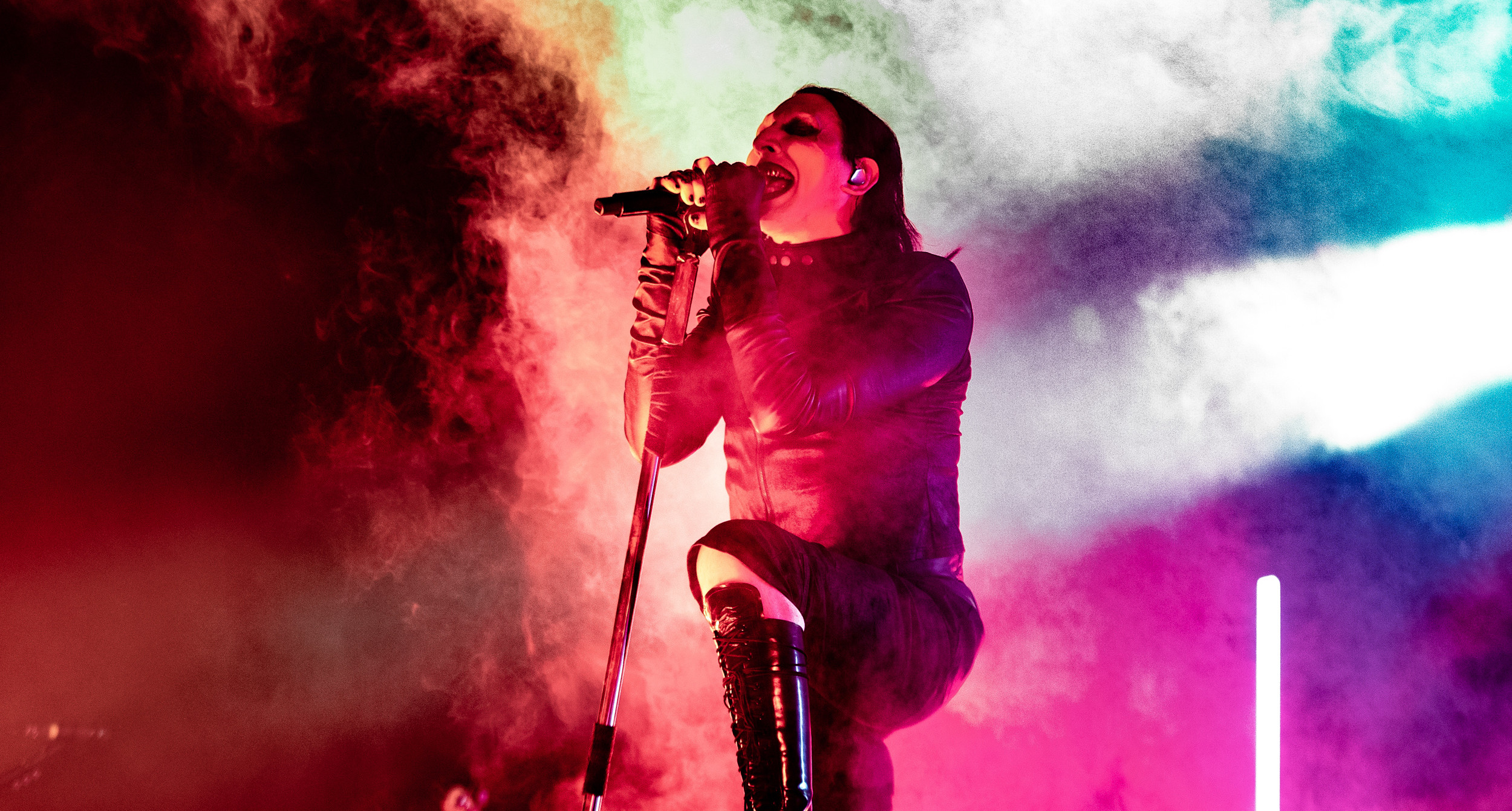 Marilyn Manson in dark attire sings passionately on stage, surrounded by vibrant red, green, and blue lights and smoke.