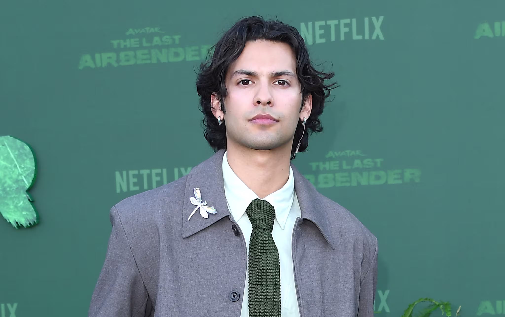 Xolo Maridueña in a gray suit with a green tie poses on a green carpet at a Netflix event for "Avatar: The Last Airbender," surrounded by plants.