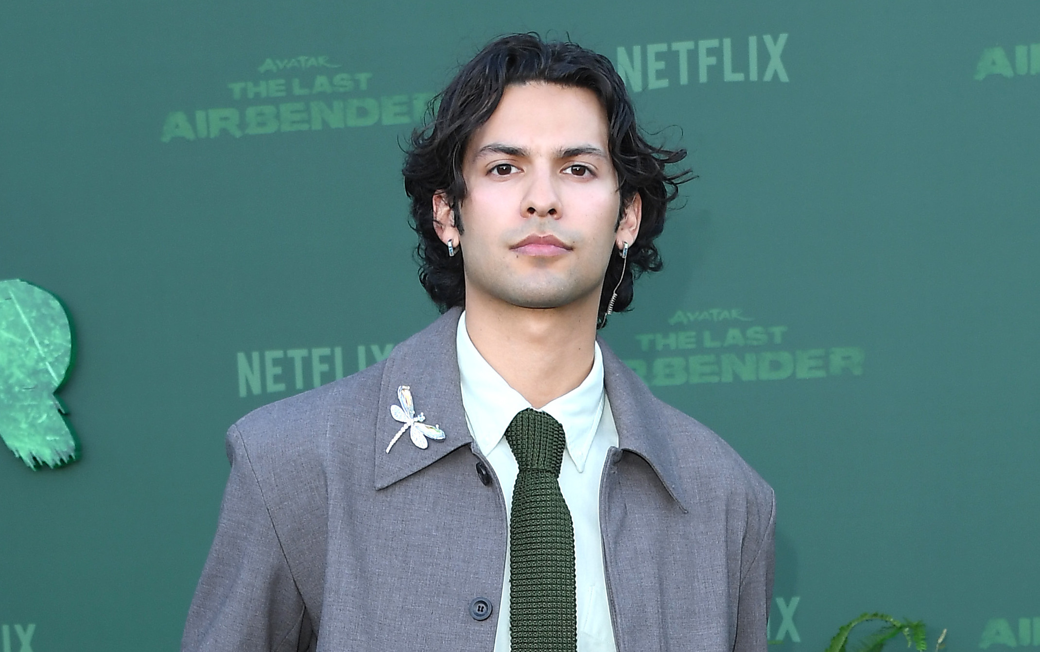 Xolo Maridueña in a gray suit with a green tie poses on a green carpet at a Netflix event for "Avatar: The Last Airbender," surrounded by plants.