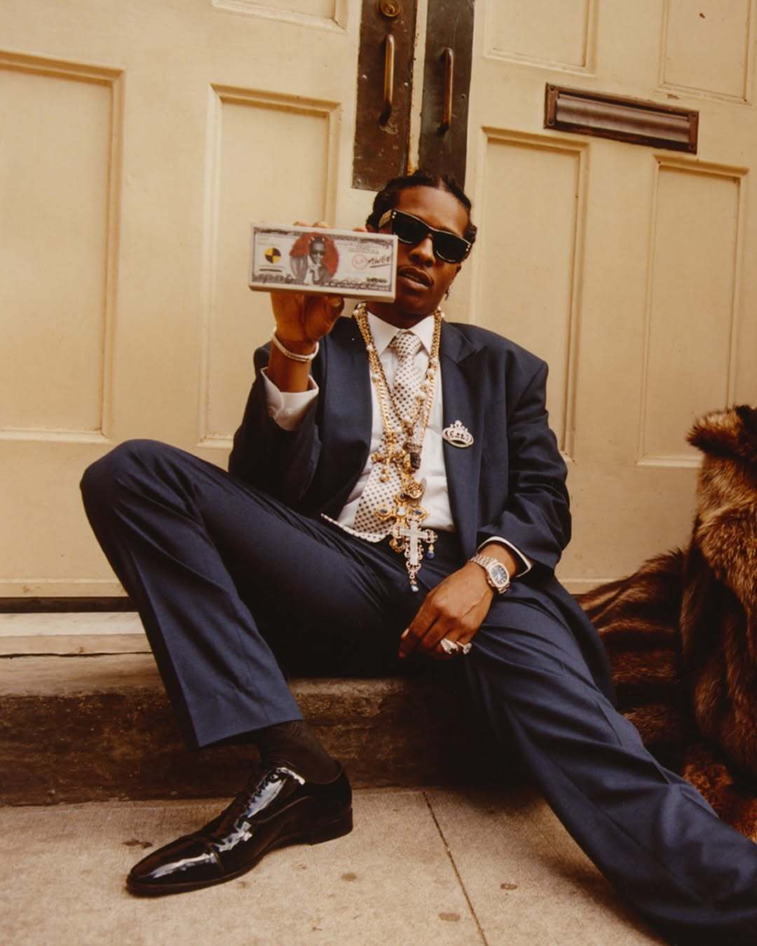 A person in a suit sits on steps, holding a bill, wearing sunglasses, and adorned with jewelry.