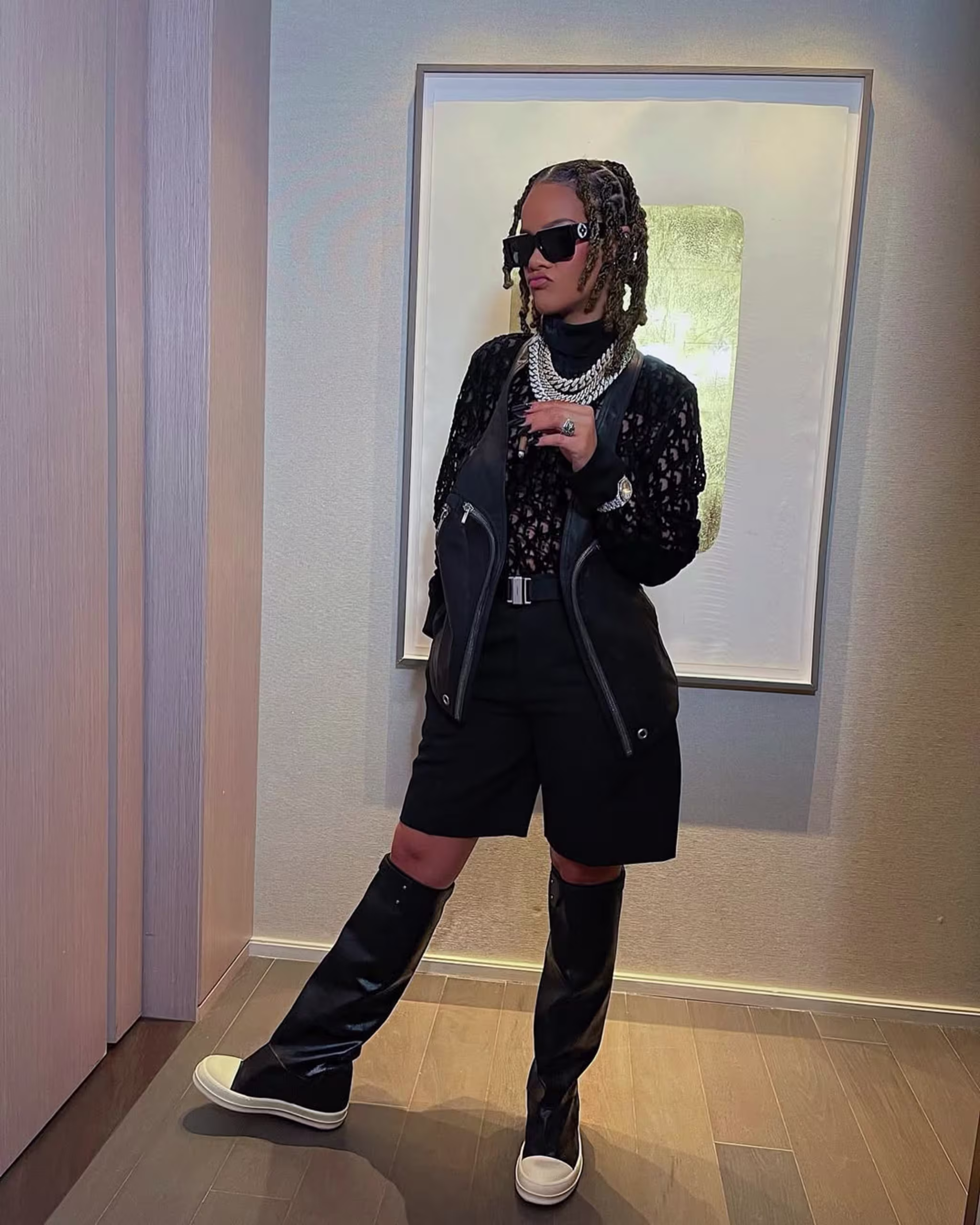 A stylish woman in sunglasses and braids, wearing a black outfit with knee-high boots, poses against a wall with framed art.
