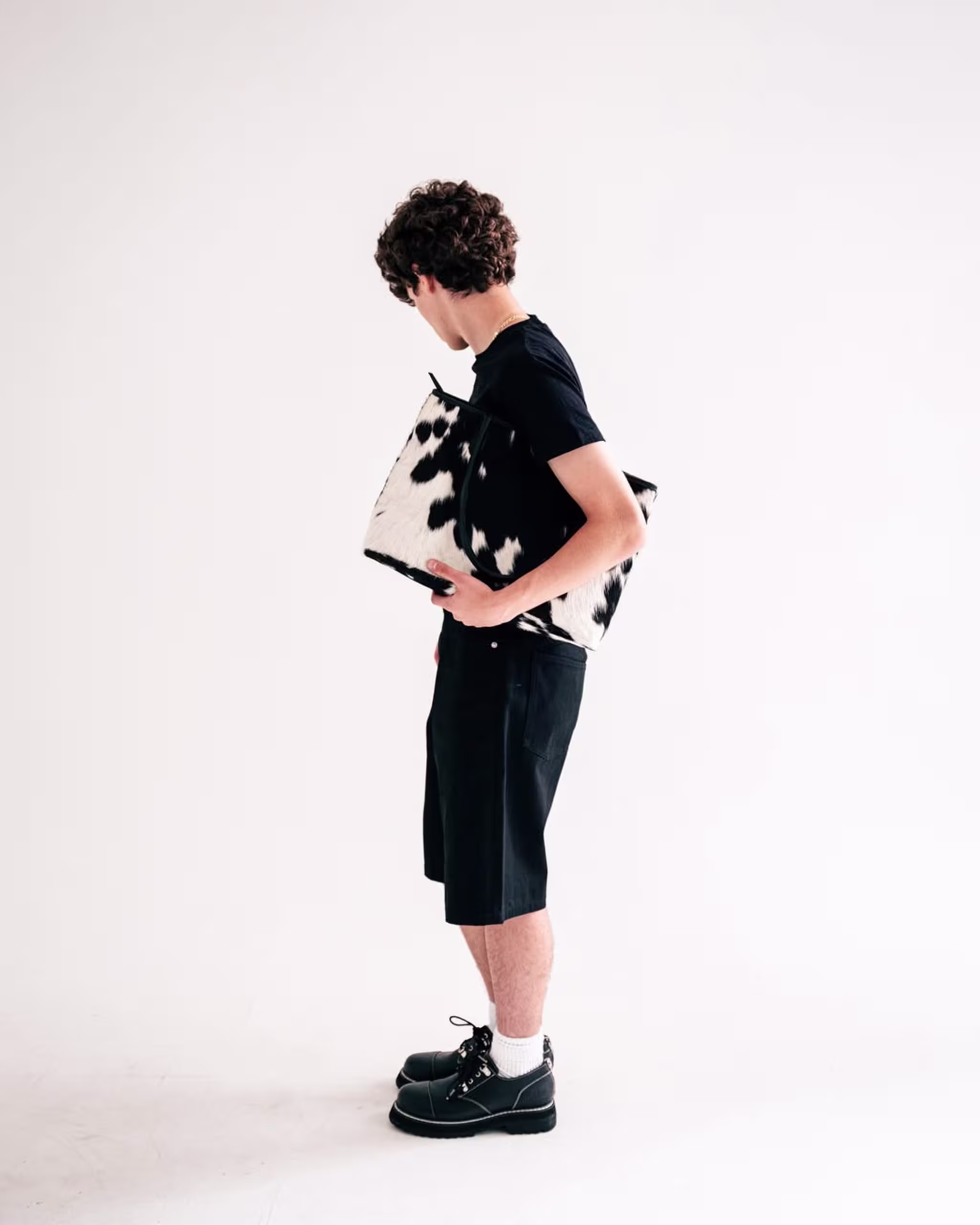 A person with curly hair, wearing a black shirt and shorts, holding a black and white patterned bag, standing against a plain background.