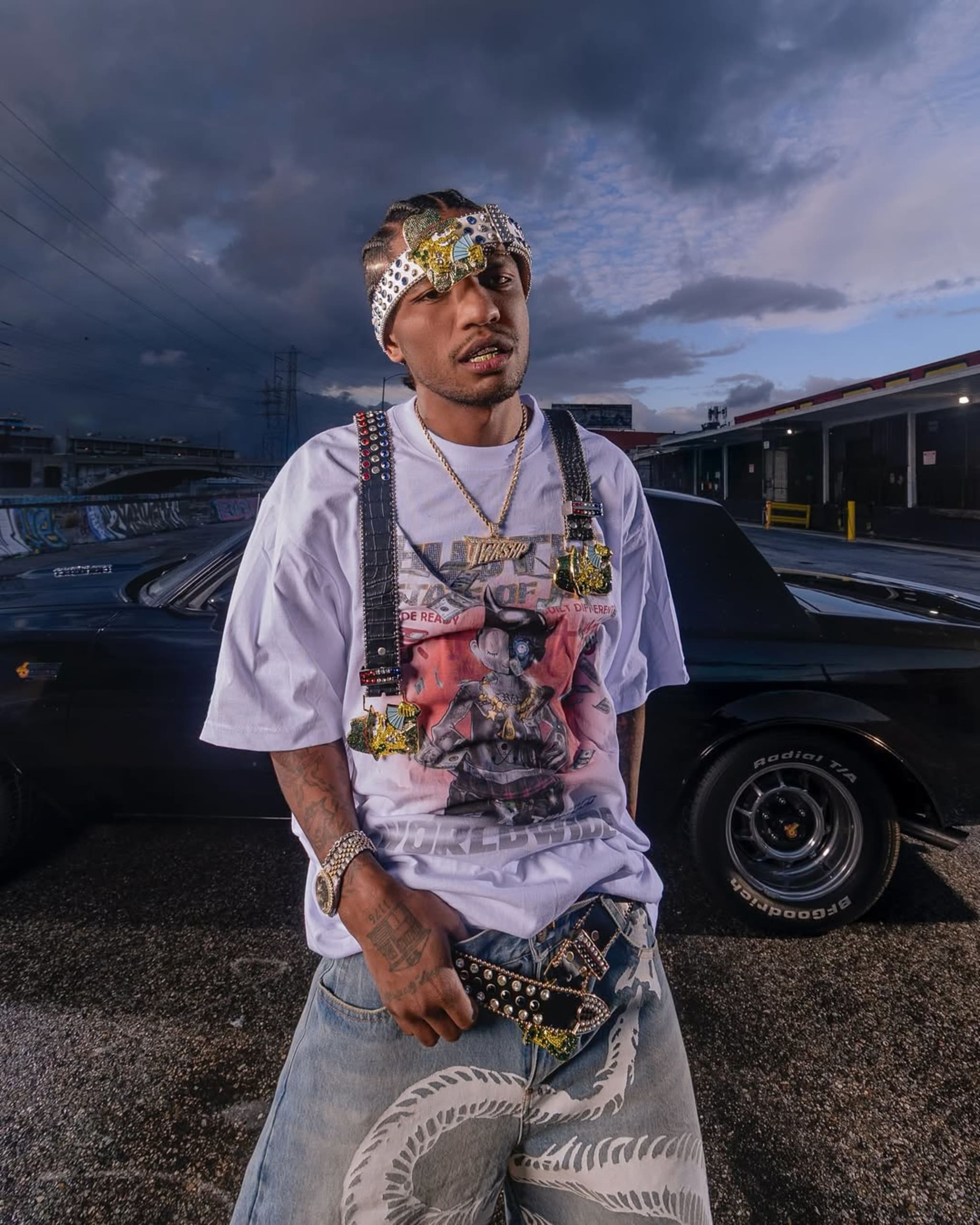 A man in urban streetwear with a graphic t-shirt, embellished accessories, and jeans stands in front of a black car under a cloudy sky.