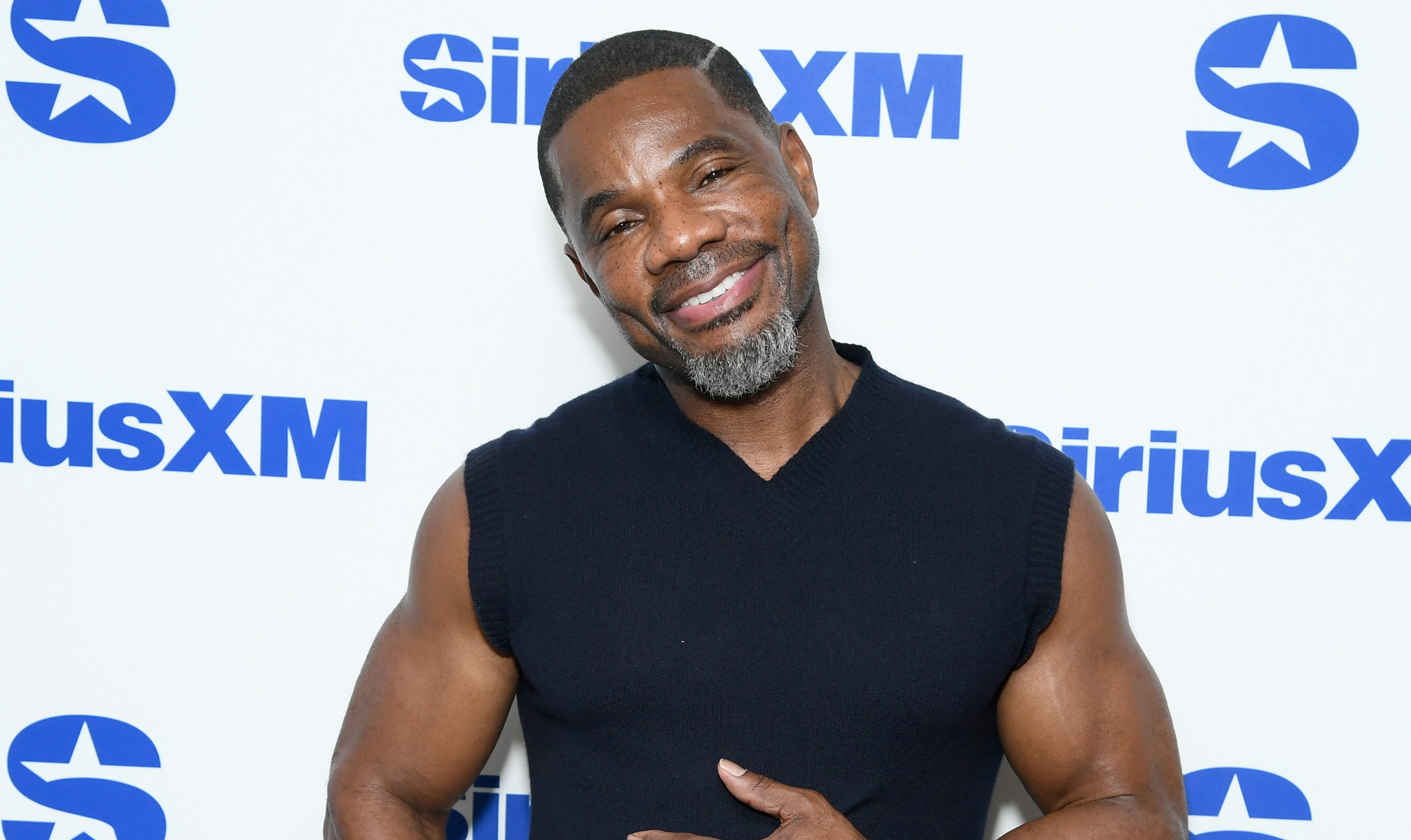 Kirk Franklin in a sleeveless sweater poses in front of a SiriusXM backdrop, smiling with one hand on his chest.