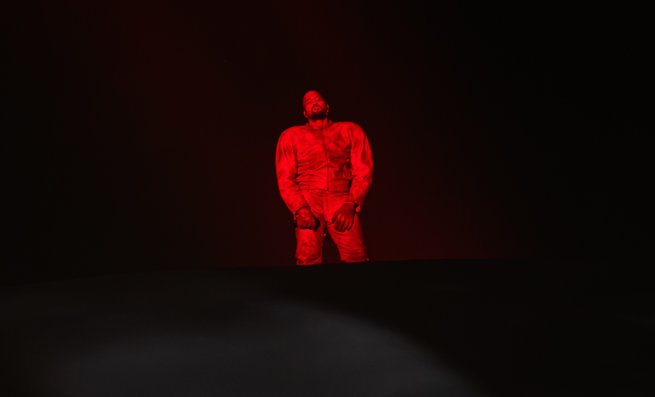 Kanye West stands on a dark stage, illuminated by red lighting, creating a dramatic effect.
