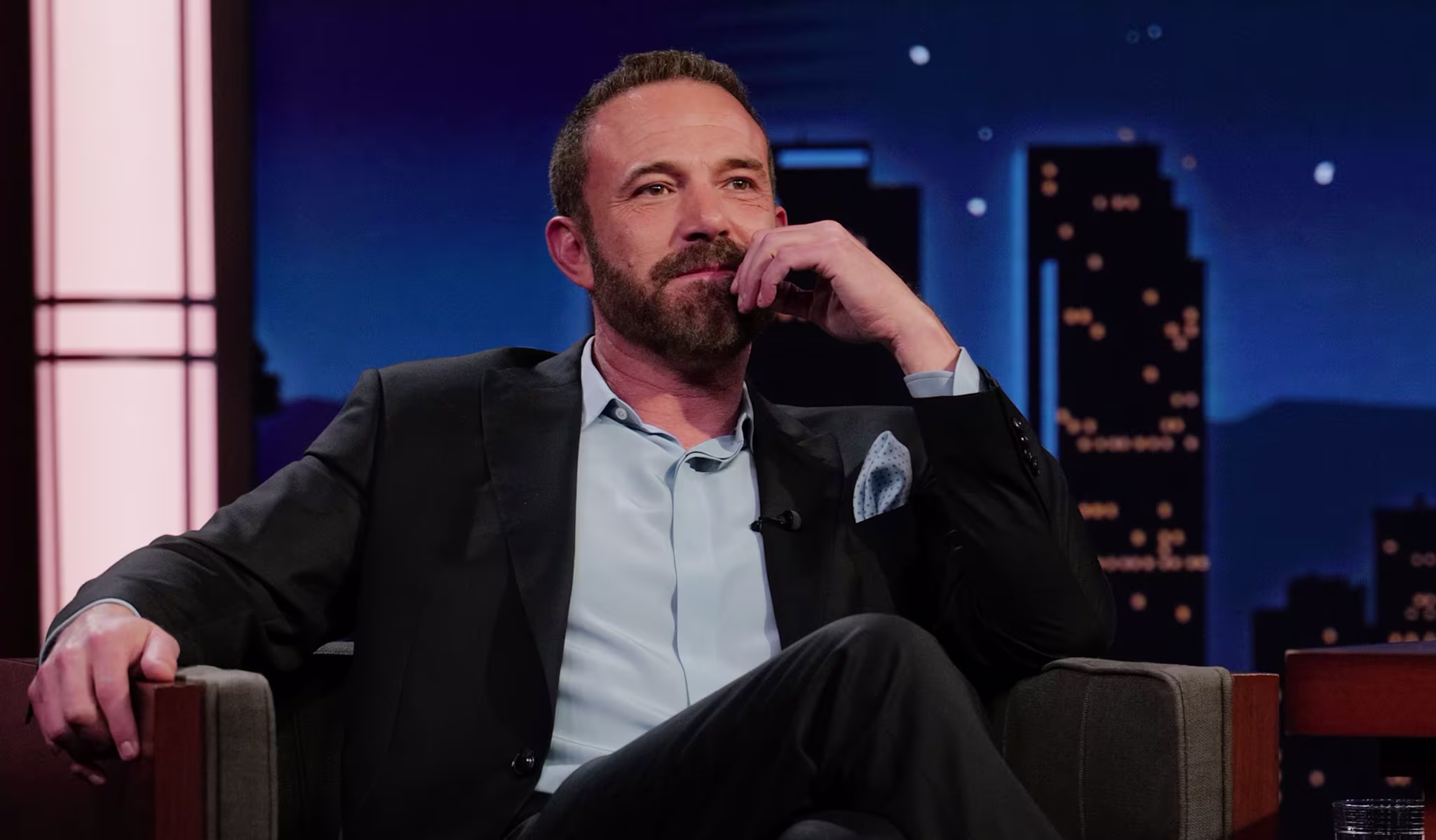 Ben Affleck in a suit sits thoughtfully on a talk show set, with a cityscape backdrop.