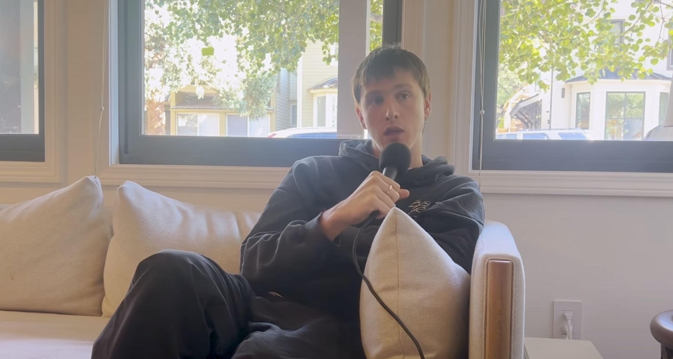 Harris Dickinson in black hoodie sitting on a couch, holding a microphone. There are windows with a view of trees and houses behind them.