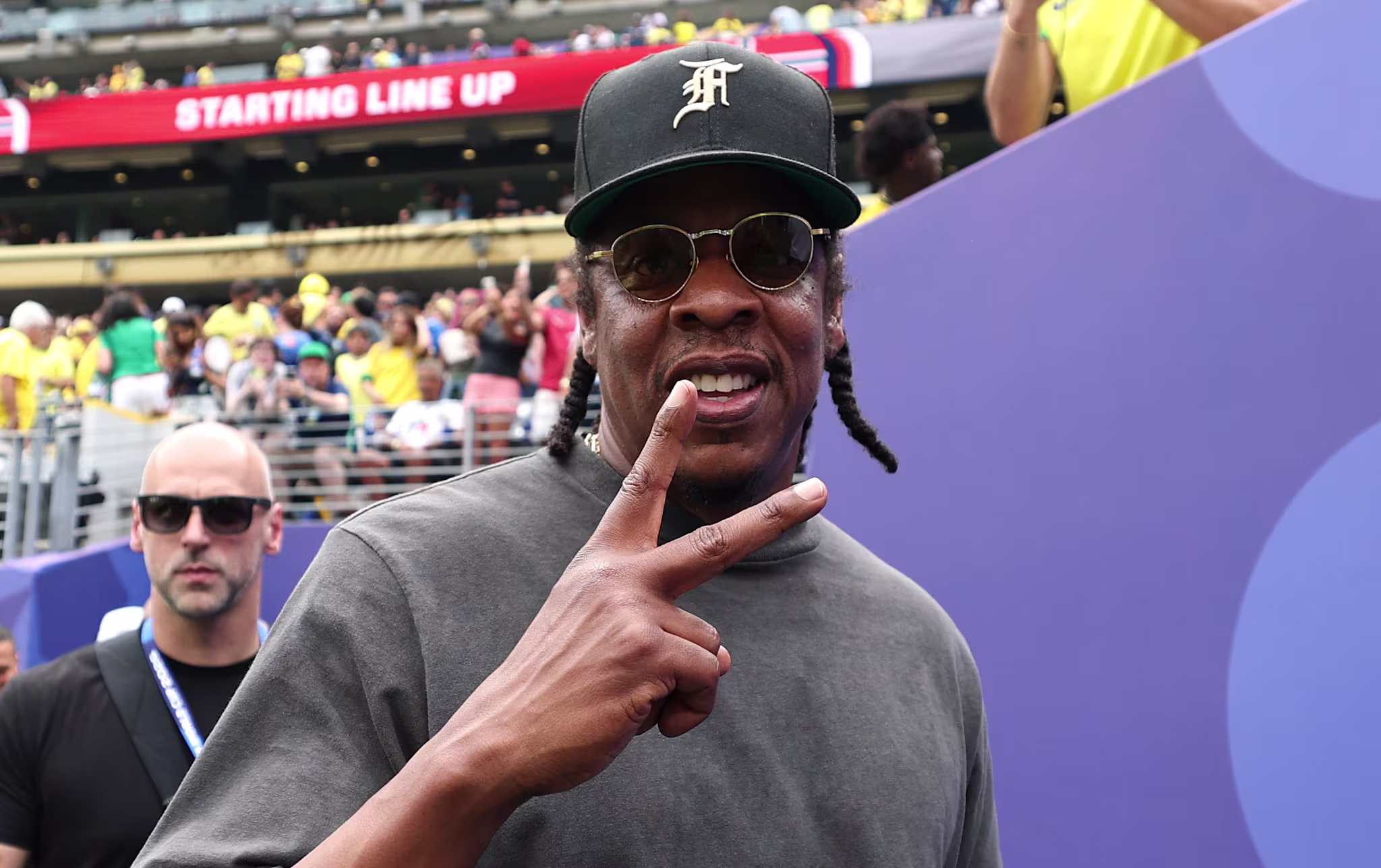 Jay-Z wearing sunglasses and a cap, flashes a peace sign at a stadium event. Crowd and security are visible in the background.