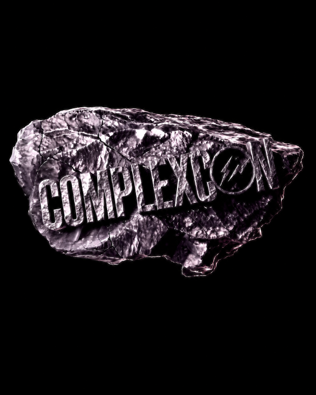Textured stone with "COMPLEXCON" engraved on it, set against a black background.