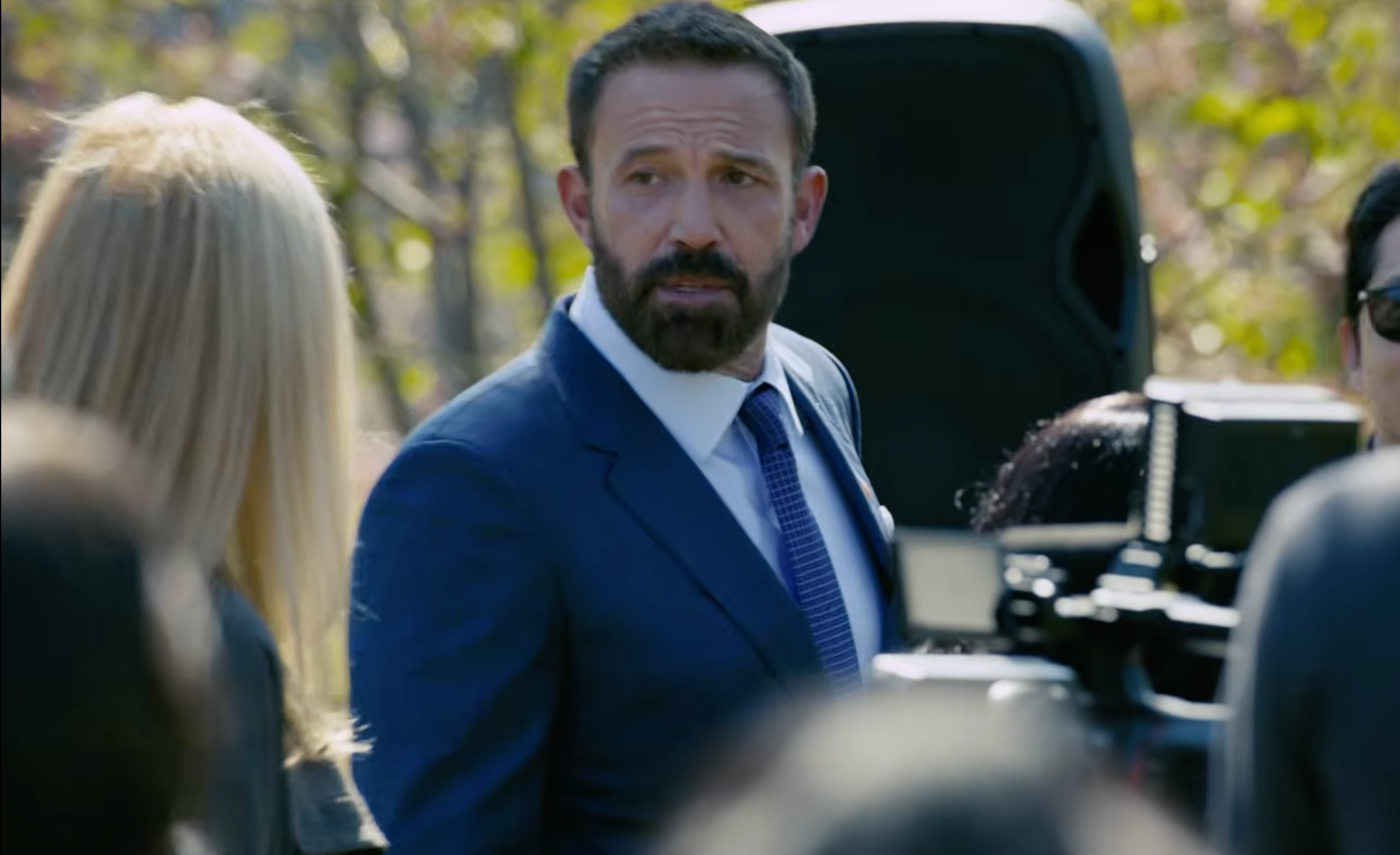 Ben Affleck in a blue suit and tie with a beard stands outdoors, looking to the side. People and equipment are around him.