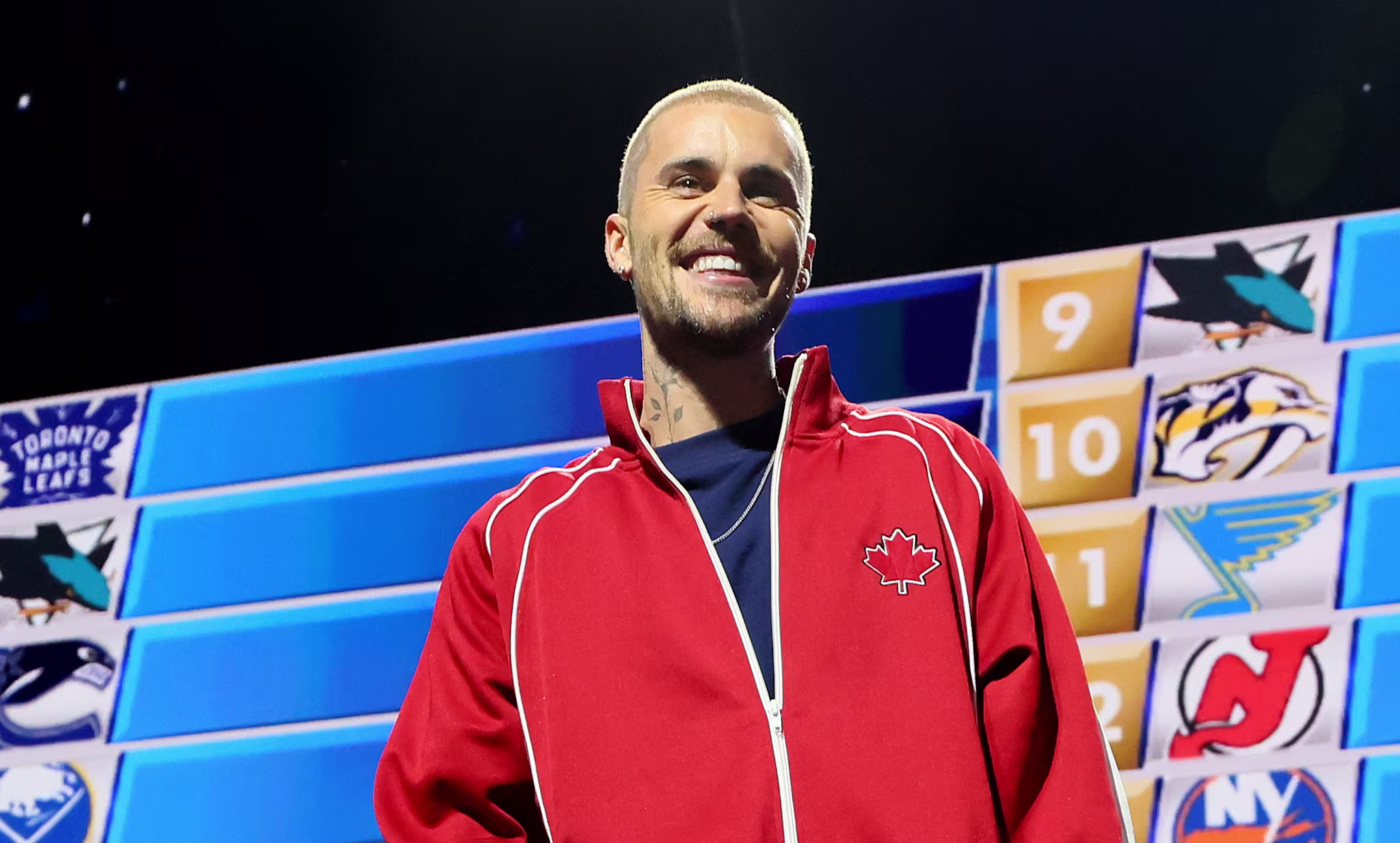 Justin Bieber in a red jacket stands on stage at an NHL event, with a draft board in the background displaying team logos.