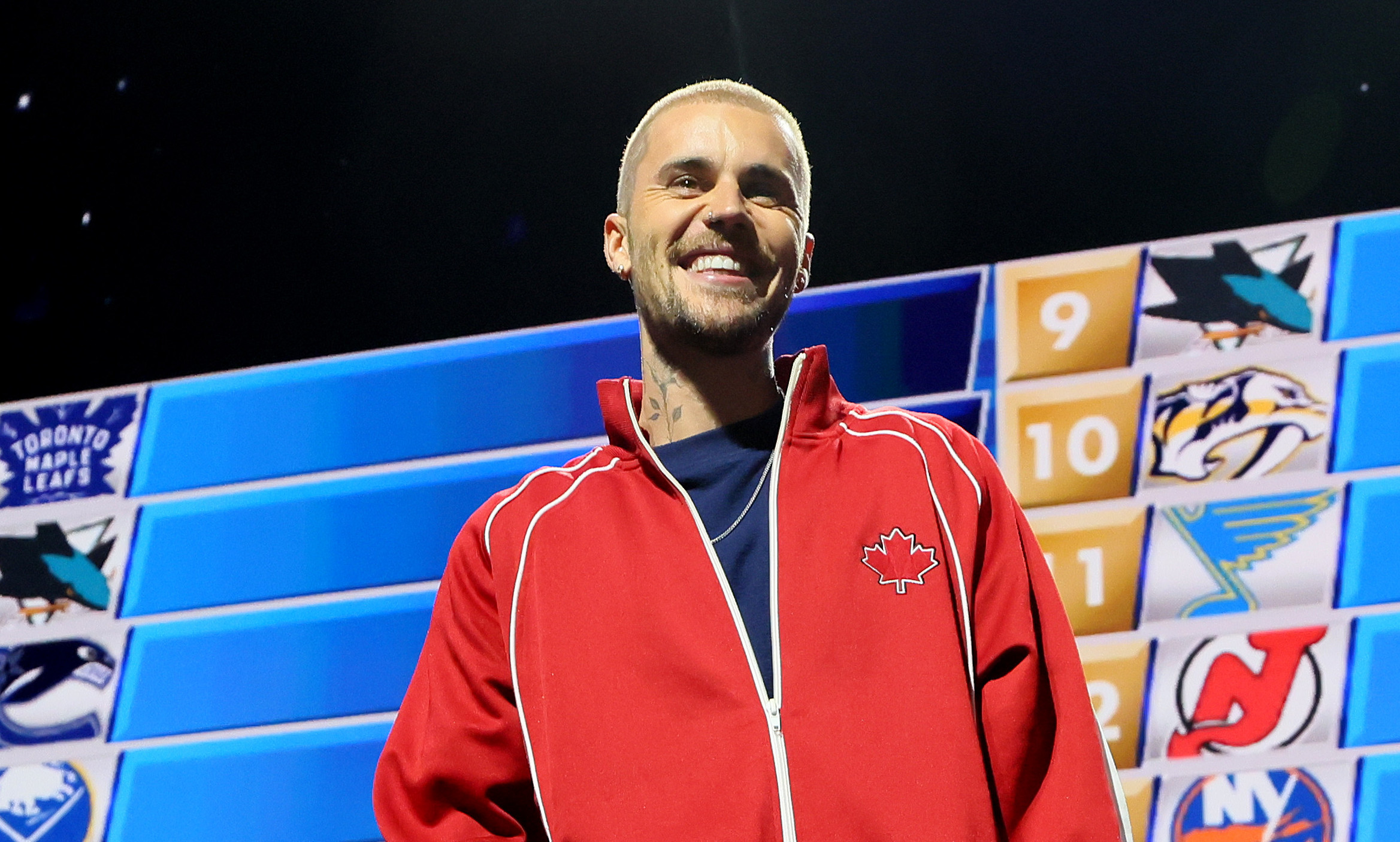 Justin Bieber in a red jacket stands on stage at an NHL event, with a draft board in the background displaying team logos.