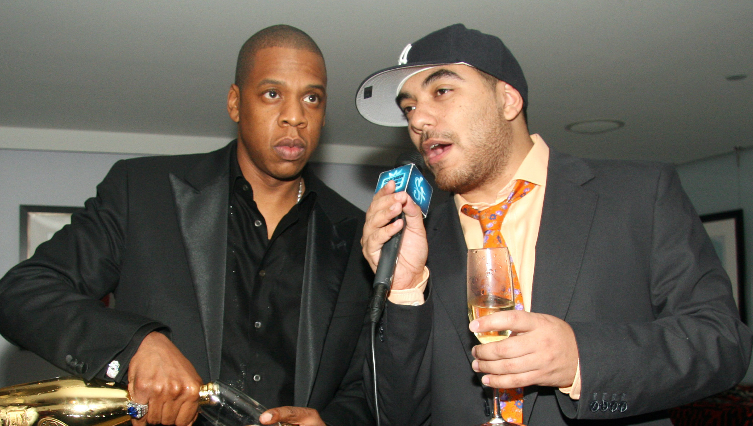 Jay-Z pours champagne while standing next to a man with a microphone, both in suits at an indoor event.