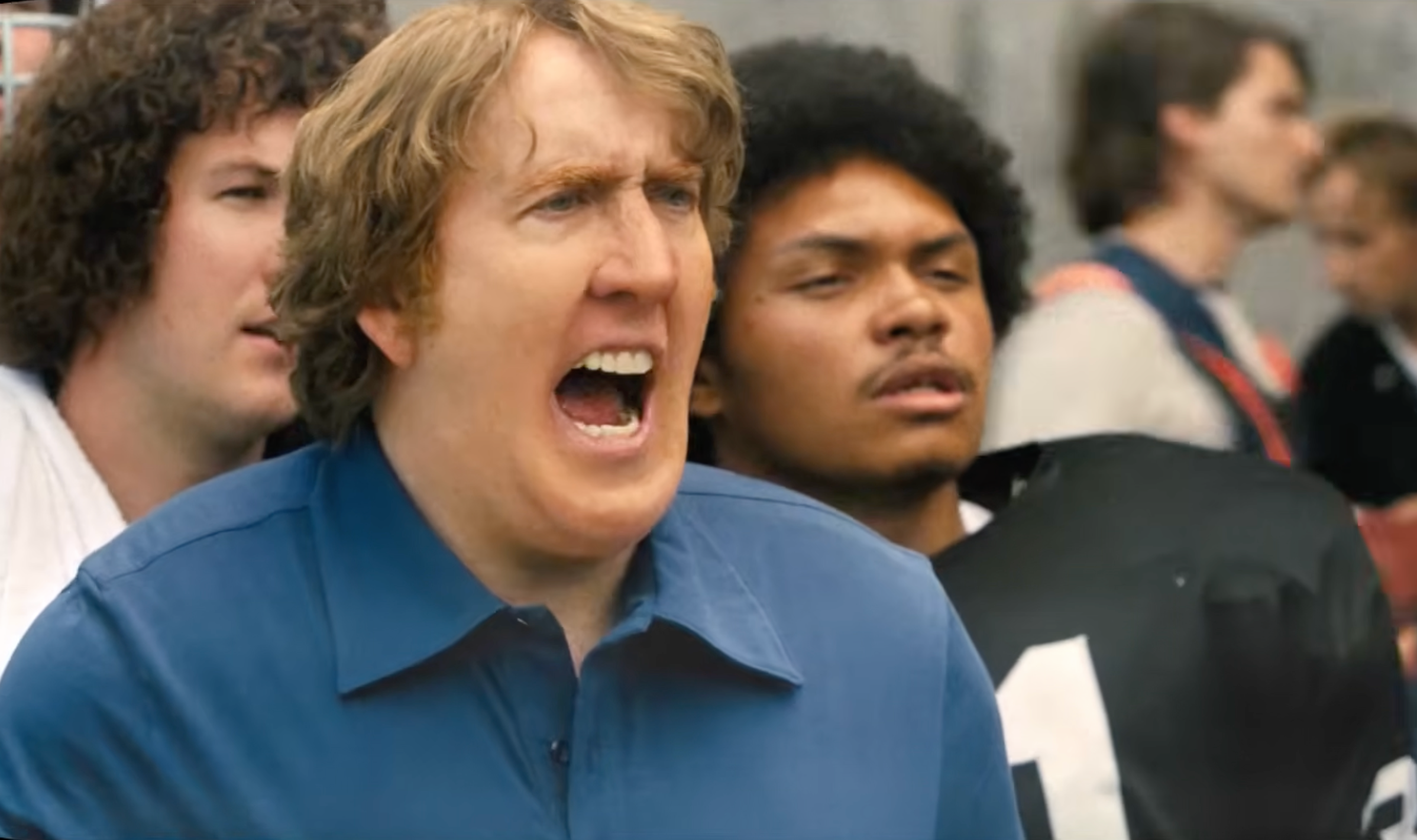 A man in a blue shirt is shouting, surrounded by others, including a person in a black sports jersey.