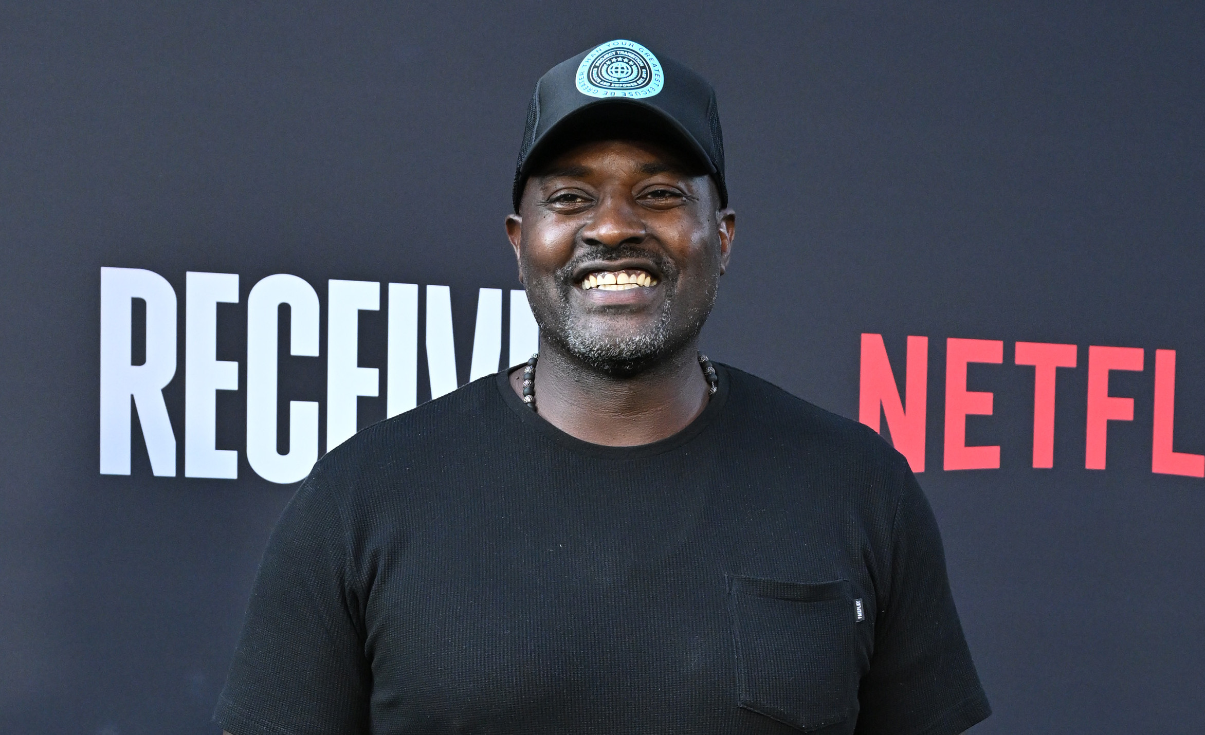Marcellus Wiley in a black shirt, jeans, and a cap stands smiling on a green carpet with a Netflix backdrop.