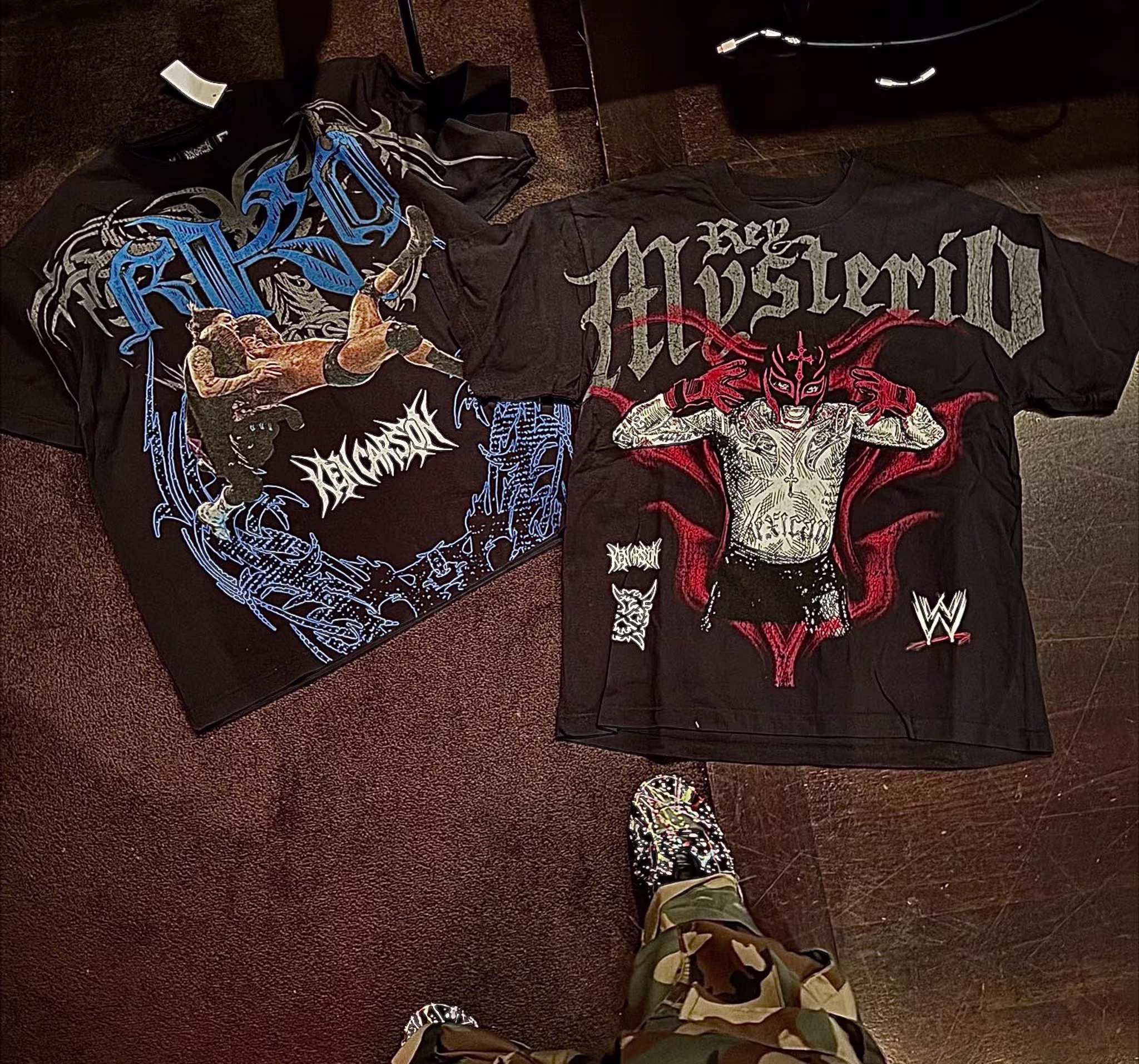 Two WWE-themed t-shirts on the floor, one featuring Rey Mysterio and the other with "RKO" and a wrestling scene. Camouflage pants and patterned shoes visible.