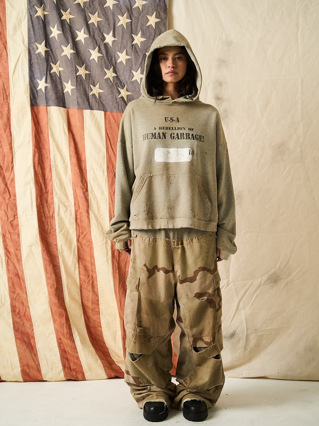 A person in a hooded sweatshirt and camo pants stands in front of a vintage American flag backdrop.
