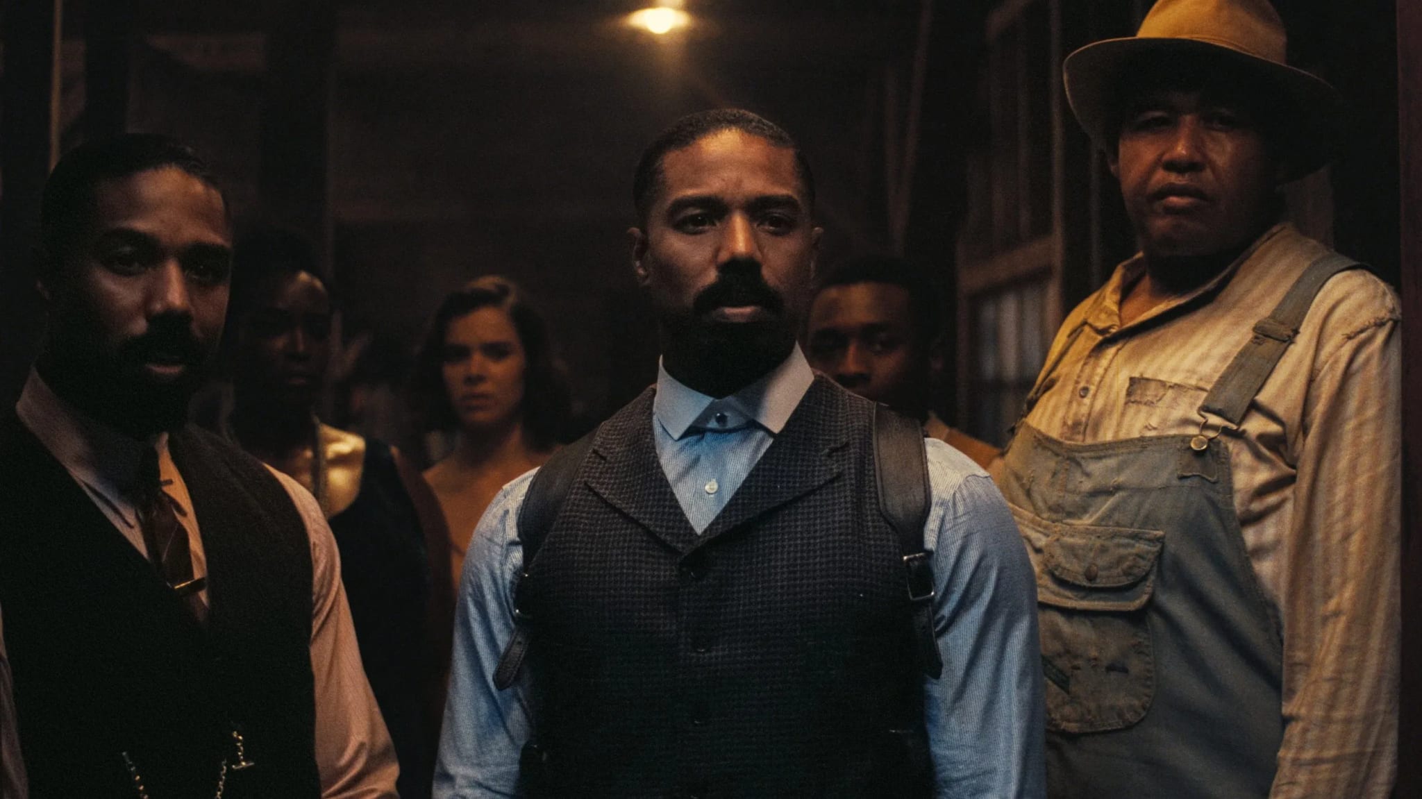 A group of people in vintage attire stands in a dimly lit room, centered around a man in a vest. The mood is serious and intense.
