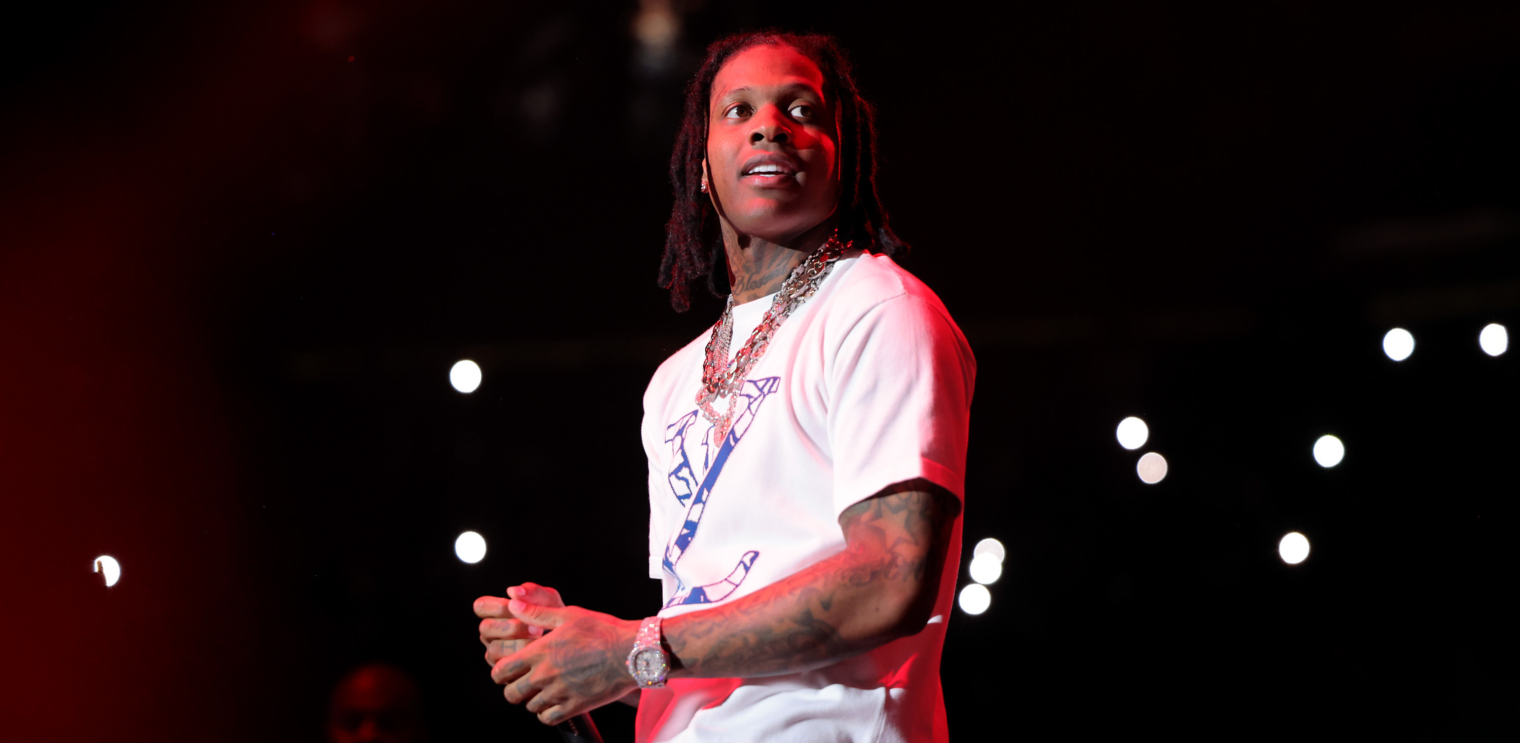 Rapper Lil Durk performing on stage, wearing a white t-shirt and jewelry, with a microphone in hand and lights in the background.