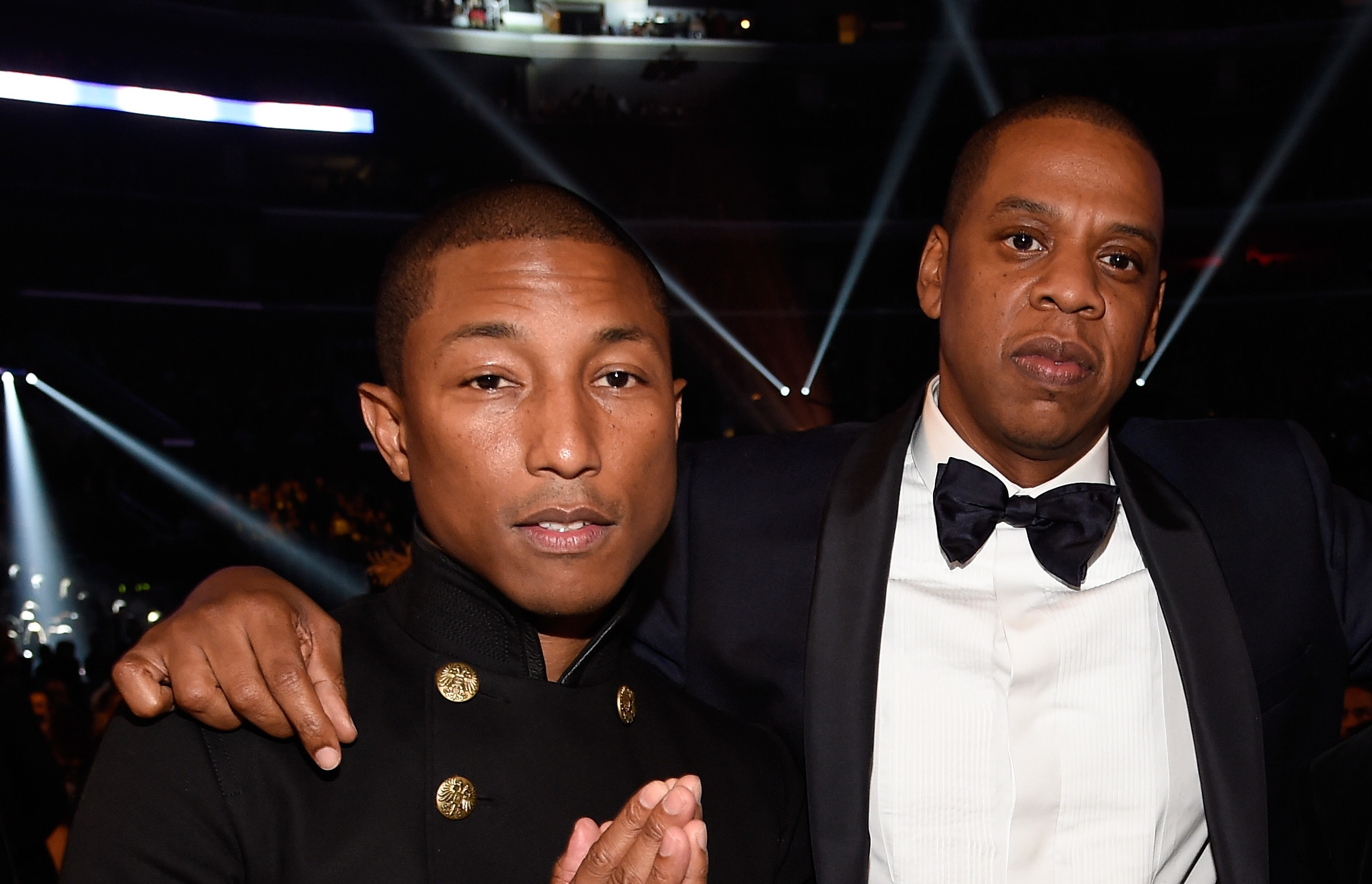 Pharrell Williams, Jay-Z, and Kanye West posing together at an event, dressed in formal attire with stage lights in the background.