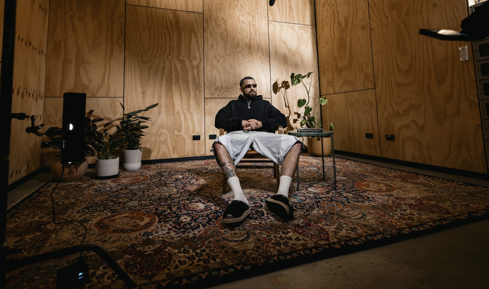 Daniel Bradshaw in sunglasses sits on a chair in a wooden room with plants, a rug, and recording equipment.