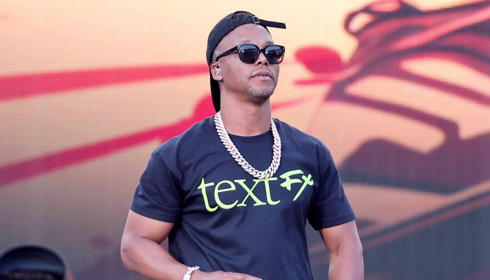 Lupe Fiasco wearing sunglasses, a black cap, and a "text FX" t-shirt performs on stage with a microphone, against a colorful backdrop.