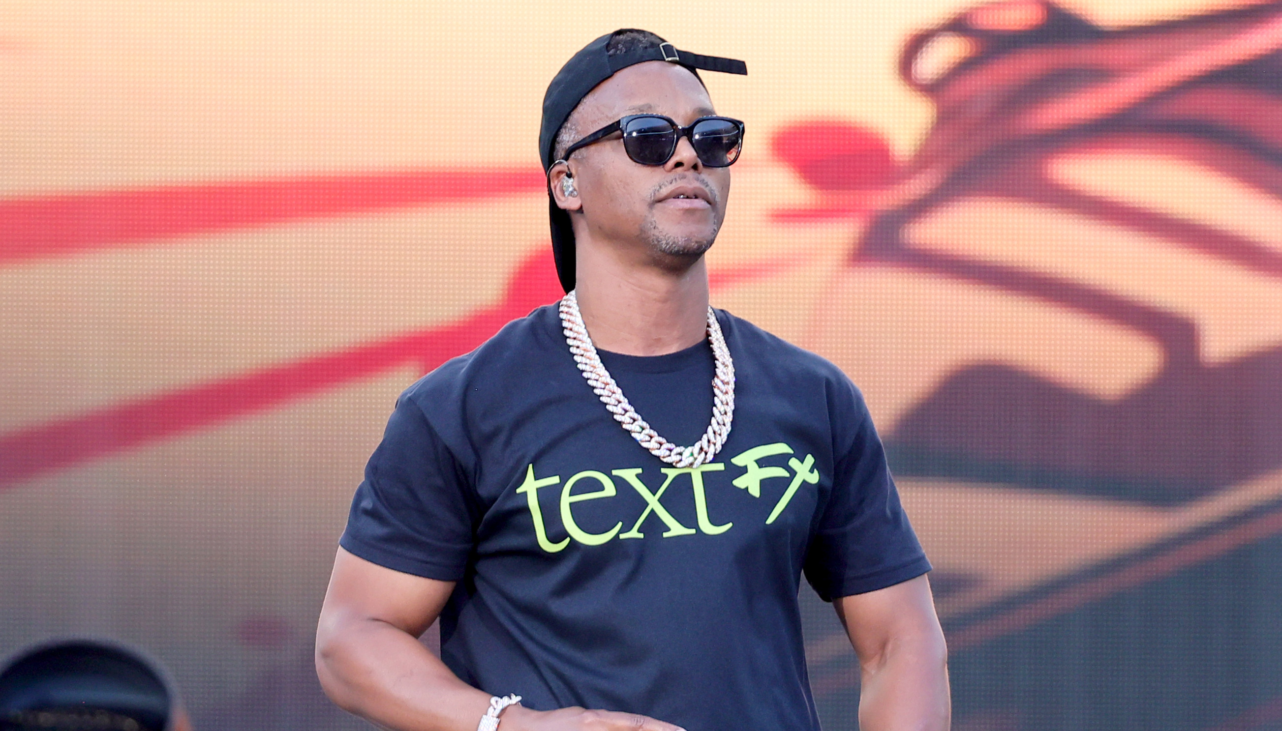 Lupe Fiasco wearing sunglasses, a black cap, and a "text FX" t-shirt performs on stage with a microphone, against a colorful backdrop.