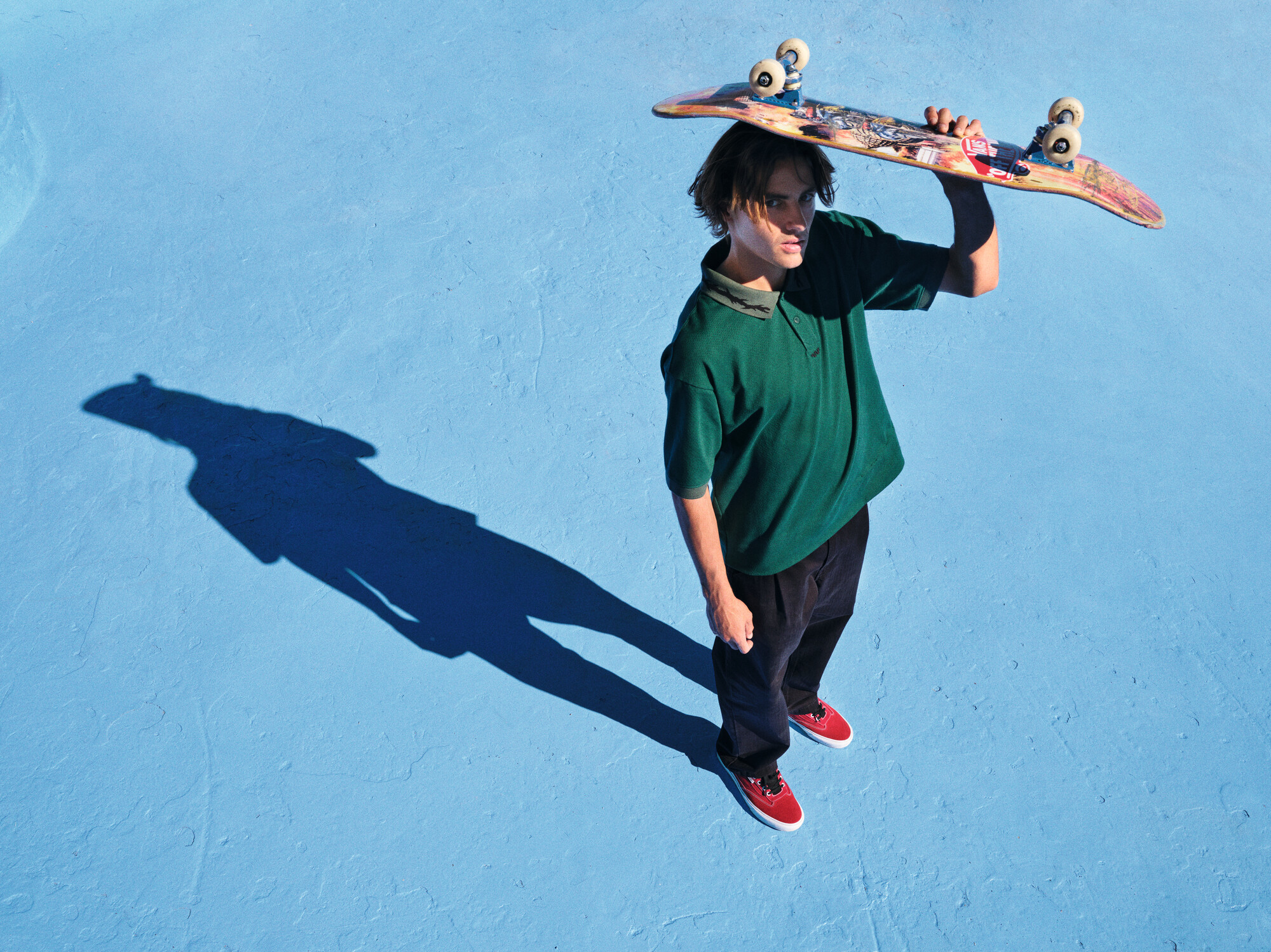 A person in a green shirt and red shoes holds a skateboard over their shoulder, standing on a blue surface with a long shadow.