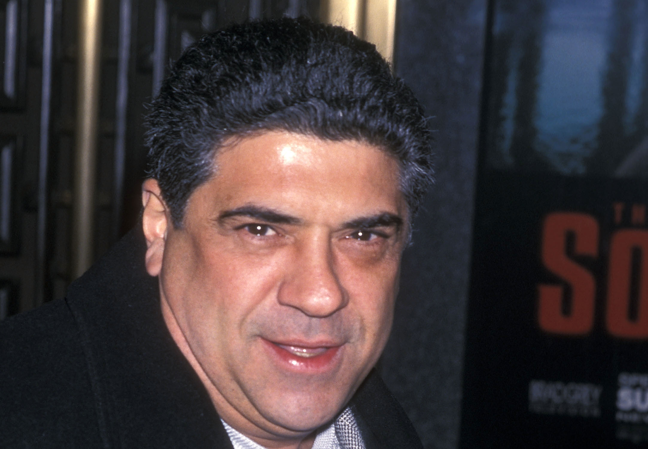 Vincent Pastore in a dark coat and sweater smiles at the camera, standing indoors with a poster in the background.