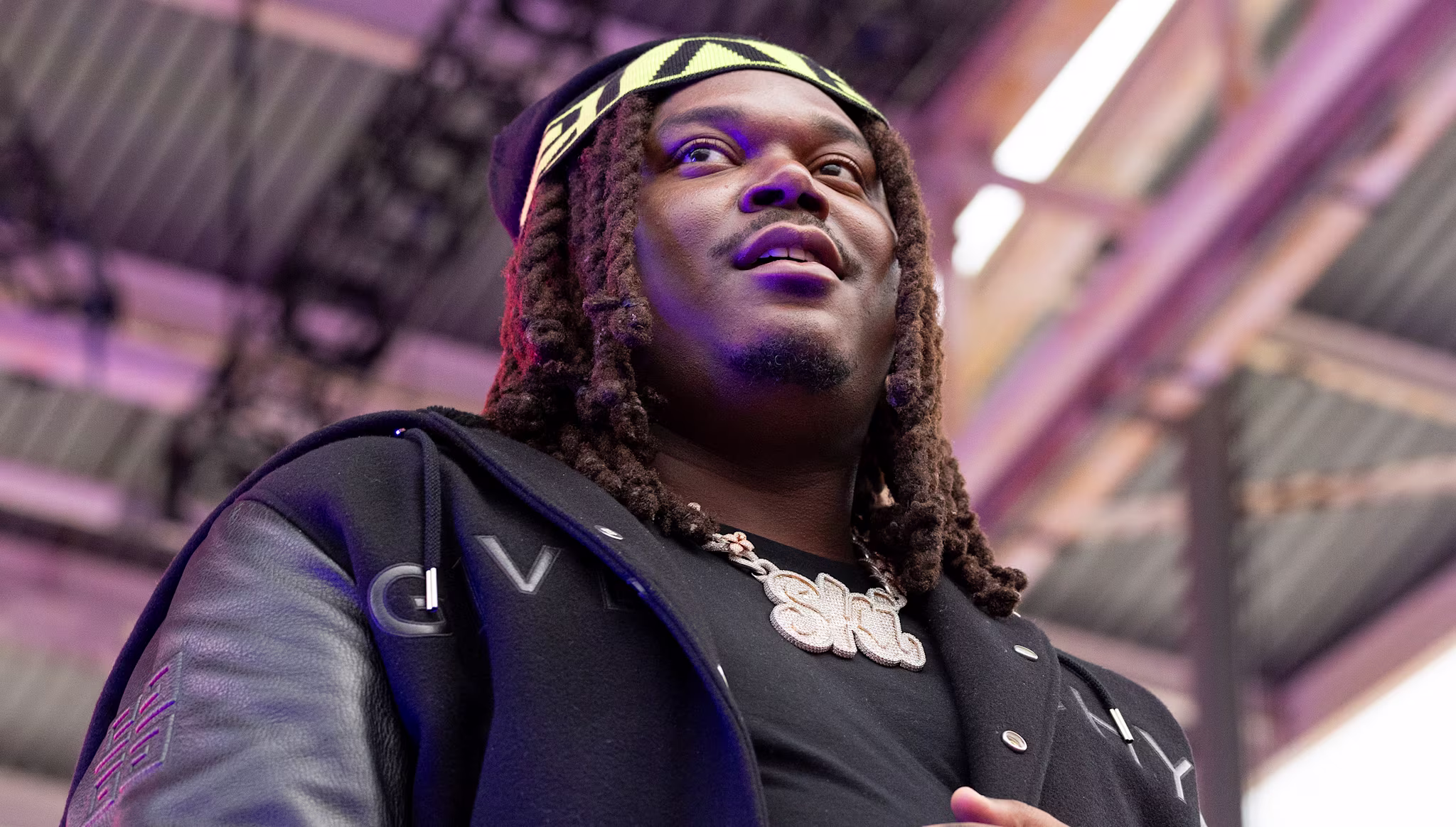 Lucki with long hair and a patterned headband, wearing a black jacket, stands under a metal structure, looking upward.
